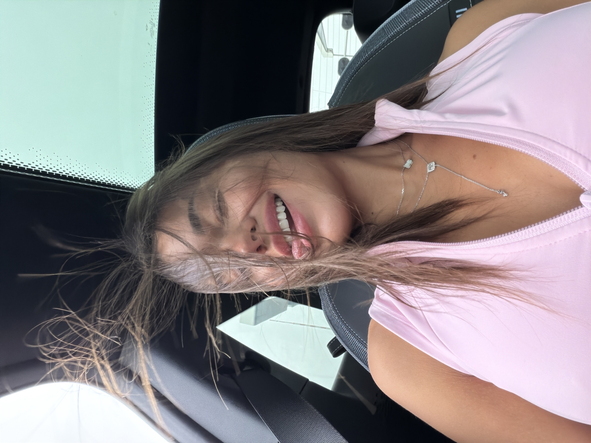 A woman with long, brown hair is captured in a selfie taken from inside a car. Her hair is blowing upwards and to the side, partially obscuring her face. She has her eyes closed, her mouth is open in a smile, and her tongue is slightly sticking out. She is wearing a light pink, sleeveless top with a zippered neckline and two delicate silver necklaces. The interior of the car is visible, including a black seat, a seatbelt, and the roof of the car, which has a sunroof. The lighting suggests it is daytime. There is no indication that the image was taken in Amsterdam, Netherlands.
