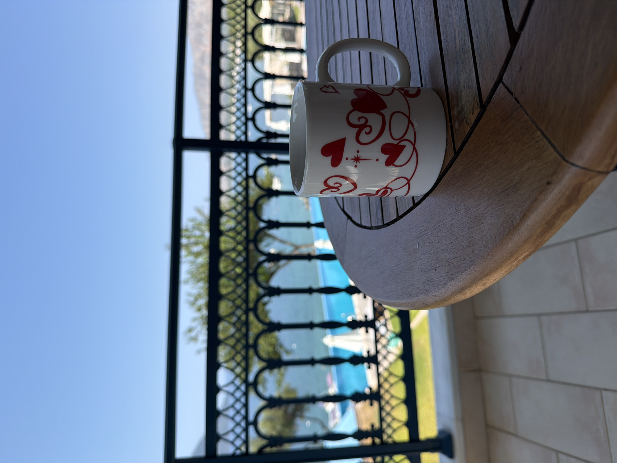 A white mug with red heart designs is placed on a wooden table on a balcony overlooking a blue swimming pool and the sea in Municipal Unit of Alyzia, Greece. The sky is clear and blue. A black metal railing separates the balcony from the view.