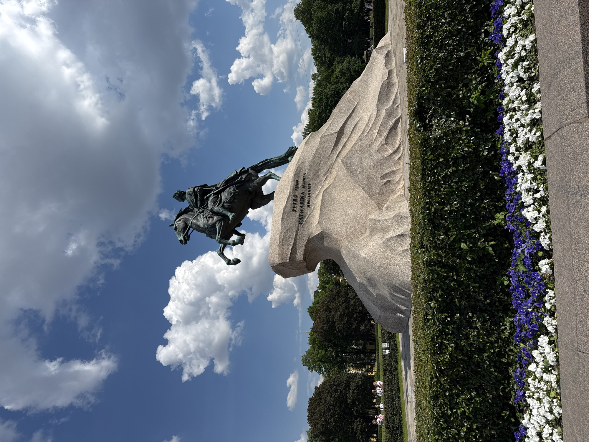 The image features the iconic Bronze Horseman equestrian statue of Peter the Great, situated in Saint Petersburg, Russia, under a bright blue sky dotted with scattered white cumulus clouds. The bronze statue, depicting Peter the Great on a rearing horse with his right arm extended, is prominently positioned on a massive, wave-like granite rock known as the Thunder Stone.
Carved into the side of the natural rock base is a Latin inscription: "PETRO PRIMO CATHARINA SECUNDA MDCCLXXXII," which translates to "To Peter the First, Catherine the Second, 1782."
In the background, a dense line of lush green trees stretches across the horizon, indicating a park or public garden. A slender, golden spire, characteristic of Saint Petersburg's skyline, is faintly visible piercing the sky above the trees in the far distance. To the right and extending into the foreground are neatly trimmed dark green hedges. Alongside these hedges, a vibrant flower bed bursts with white flowers interspersed with clusters of purple blooms.
The scene is illuminated by bright natural sunlight, suggesting a clear and pleasant day. A few small, indistinct figures can be observed in the far background near the treeline, indicating the presence of visitors enjoying the monument and its surroundings. The image captures a majestic and serene view of this historical landmark.