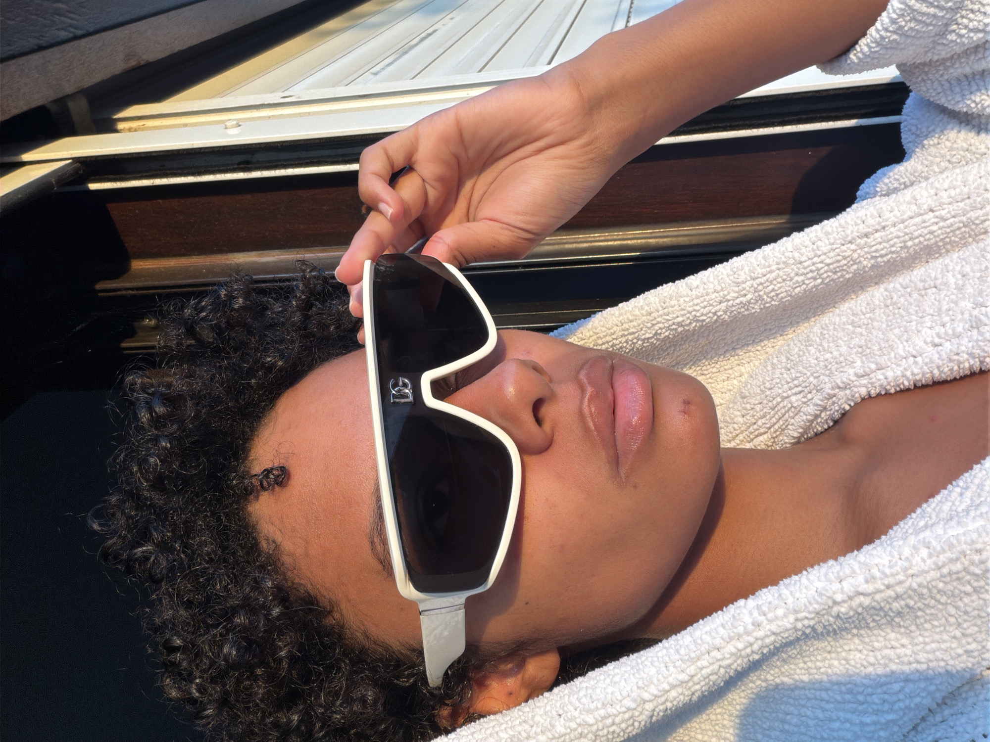 A young person with dark, curly hair and fair skin is wearing oversized, white, wrap-around sunglasses with black lenses and a silver logo on the frame. They are also wearing a white, terrycloth robe. The person is looking towards the right side of the frame, with their right hand raised to adjust the sunglasses. The background consists of a dark surface on the left and a window frame with blinds on the right, illuminated by warm, natural light, suggesting an outdoor or well-lit interior setting. The scene appears to be captured during golden hour.
The specific location in Champs-sur-Marne, France, is unknown from the image content alone.
