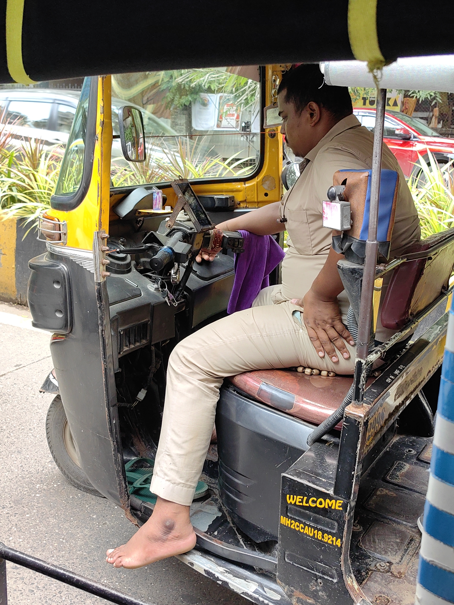A man, likely an auto-rickshaw driver, is seated inside a yellow auto-rickshaw. He is wearing a light-colored uniform shirt and pants. His left foot is bare and resting on the edge of the auto-rickshaw, revealing a dark bruise or mark on his ankle. A purple cloth is draped over the steering column. The auto-rickshaw appears to be parked on a street in Mumbai, India, as indicated by the license plate MH2CCA U18.9214. In the background, there are green plants and the side of a red car. The overall scene suggests a snapshot of daily life in Mumbai.