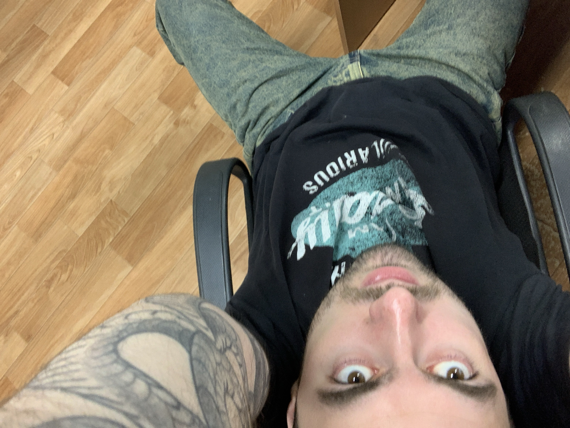 An indoor, low-angle selfie captures a young man, likely in his 20s or 30s, looking directly at the camera with wide, surprised eyes and slightly parted lips. He is positioned as if reclining or lying back in a chair, with his head tilted, causing the image to appear rotated with the floorboards running diagonally. His face is central and dominant in the frame, indicating a very close-up shot.
The man has a short beard or stubble and brown eyes. On his left arm and shoulder, a large, intricate tattoo is visible, primarily in black and gray tones, featuring elaborate, possibly wing-like or abstract designs. He is wearing a black t-shirt with a faded teal and white graphic design across the chest. The visible part of the text on his shirt, appearing upside down from this perspective, reads "LARIOUS," suggesting it is part of a larger word like "HILARIOUS." He is also wearing faded blue or acid-wash jeans, parts of which are visible in the upper half of the image.
He appears to be sitting or lying in a black office chair, with the armrests visible on either side of his torso. The background consists of a light-colored wooden floor with planks laid diagonally, and a dark brown object, possibly furniture or a box, is partially visible in the upper right corner. The lighting is soft and even, typical of an indoor environment, with no specific time of day or weather cues visible. The overall mood is playful or surprised due to his expression. This image was taken in Chaikovsky, Russia.