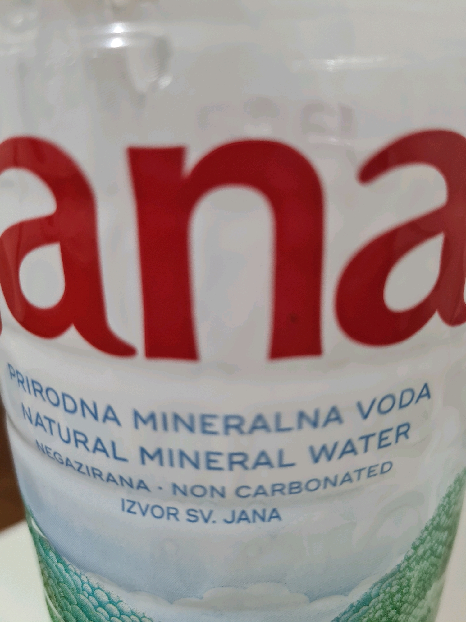 The image shows a close-up of a plastic bottle of "Jana" natural mineral water. The brand name "Jana" is prominently displayed in large red lettering across the top of the bottle. Below the brand name, in smaller blue lettering, are the words "PRIRODNA MINERALNA VODA", "NATURAL MINERAL WATER", and "NEGAZIRANA - NON CARBONATED". The text "IZVOR SV. JANA" is also visible. The bottle is white with a textured pattern of green and white at the bottom, resembling stylized leaves or scales. The background is blurred and appears to be a neutral indoor environment. The image does not contain any information about the city of Rijeka, Croatia.