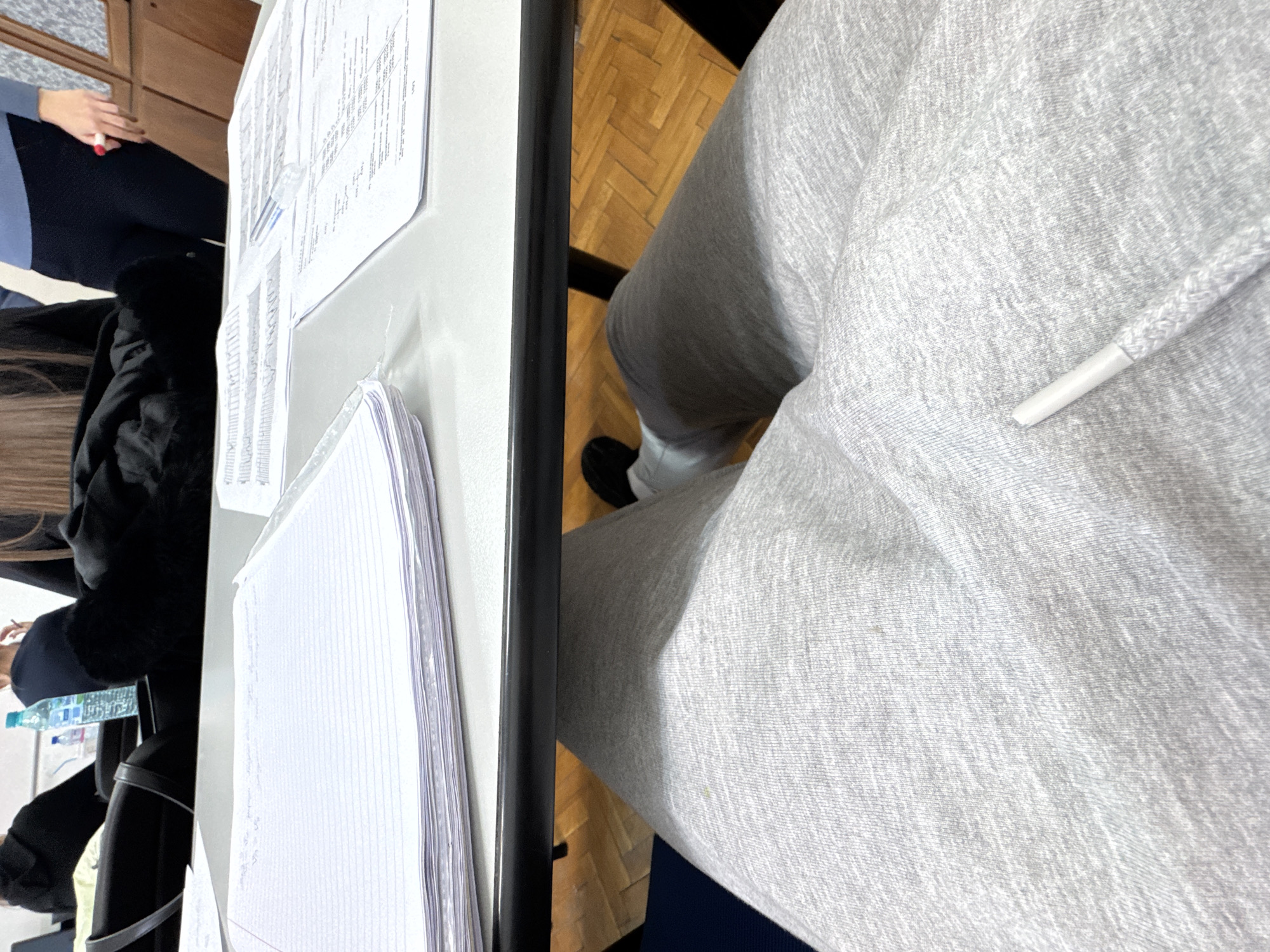 A photo taken from an above perspective shows a desk setting. On the left side of the frame is a table with various papers and notebooks stacked. A person wearing a black jacket sits near the table, while another person with blue sleeves is partially visible. A water bottle can be seen on the table. To the right is a person wearing gray sweatpants. Part of a wooden floor is visible beneath the table and legs. The location is a room somewhere in Sofia, Bulgaria.