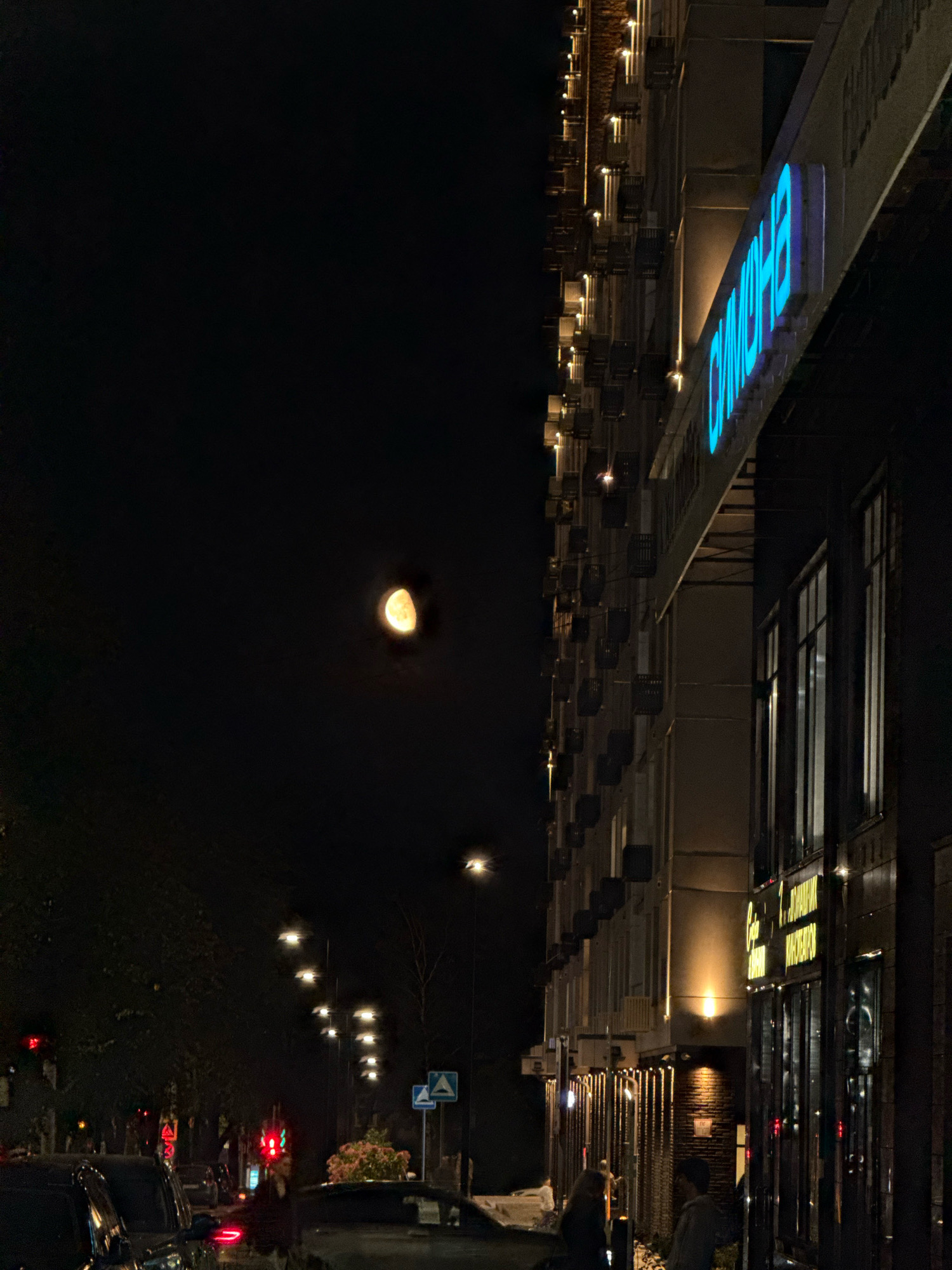 In Nizhny Novgorod, Russia, a nighttime street scene unfolds with a modern building illuminated by streetlights and neon signage. The building's facade features rows of windows and balconies, with a prominent blue neon sign that appears to spell out "ОЛИМПИЯ" (Olympia). Across the street, a line of streetlights guides the way along a dark road where a few cars are visible, including a black car in the foreground with its taillights illuminated. The moon, partially visible and glowing orange, hangs in the dark sky. The scene suggests a vibrant urban environment at night.