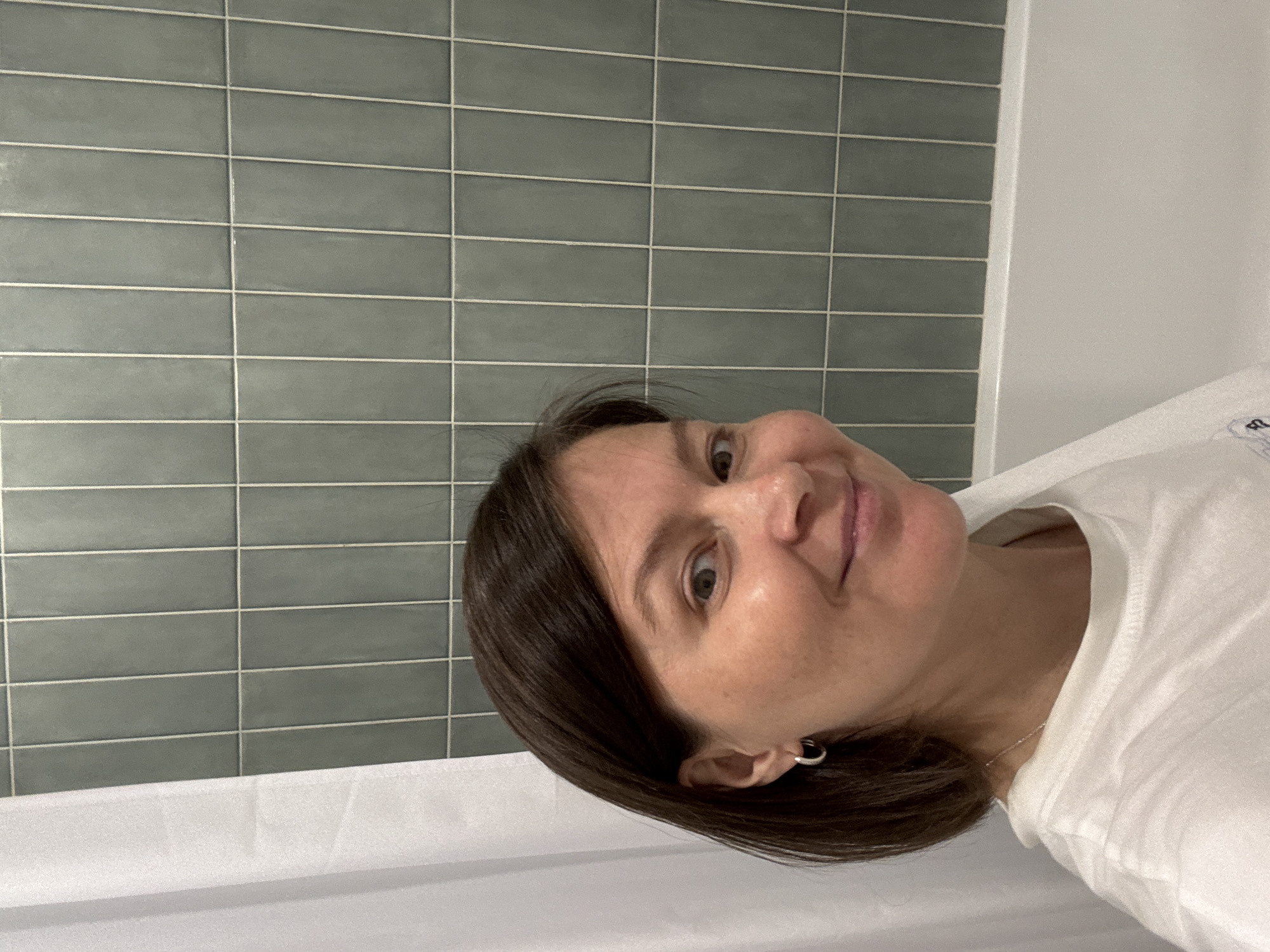 A woman is taking a selfie in a bathroom. She has short, dark brown hair and is wearing a white t-shirt and silver hoop earrings. She is smiling at the camera. The background shows a tiled wall and a white bathtub. The image does not contain any information that would allow for its location to be identified, including in Izhevsk, Russia.
