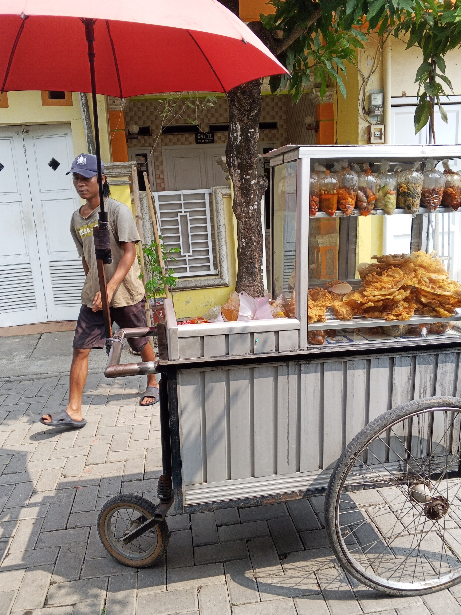 In Ciruas, Indonesia, a street food vendor stands near his cart under a red umbrella. The vendor, wearing a grey t-shirt and dark shorts, holds a pole attached to the cart. The cart displays various fried foods in clear plastic bags on shelves. The scene is set on a paved sidewalk in front of a building with white doors and yellow accents. A large wheel is visible on the right side of the cart.