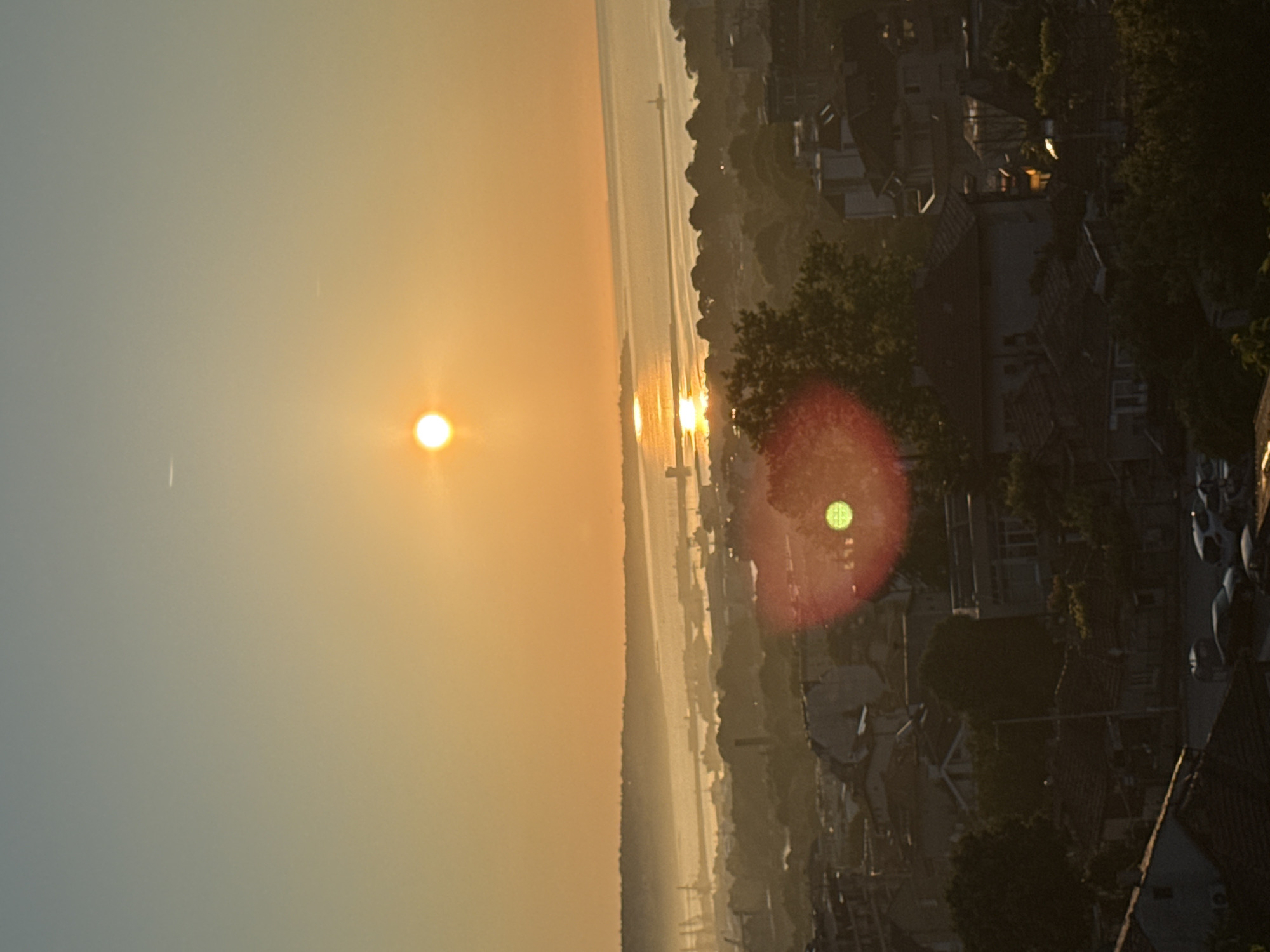 The image captures a panoramic view of a city at sunset. The sun, a bright orange orb, hangs low in the sky, casting a warm glow over the scene. Its reflection shimmers on the calm waters of a bay or large river in the mid-ground. A hazy silhouette of landmasses and industrial structures, possibly a port with cranes and ships, is visible across the water. The foreground is dominated by the rooftops of residential buildings, characterized by terracotta tiles. Trees are scattered throughout the urban landscape. A prominent lens flare, appearing as a reddish-orange circle with a green center, is present in the middle-right portion of the image. Several vehicles are parked on a street in the lower left of the frame, and a light fixture is visible on a pole in the lower right. The sky transitions from a pale blue at the top to a warm orange hue near the horizon, with faint white contrails from aircraft visible. The overall impression is a serene urban sunset overlooking a body of water and industrial area.