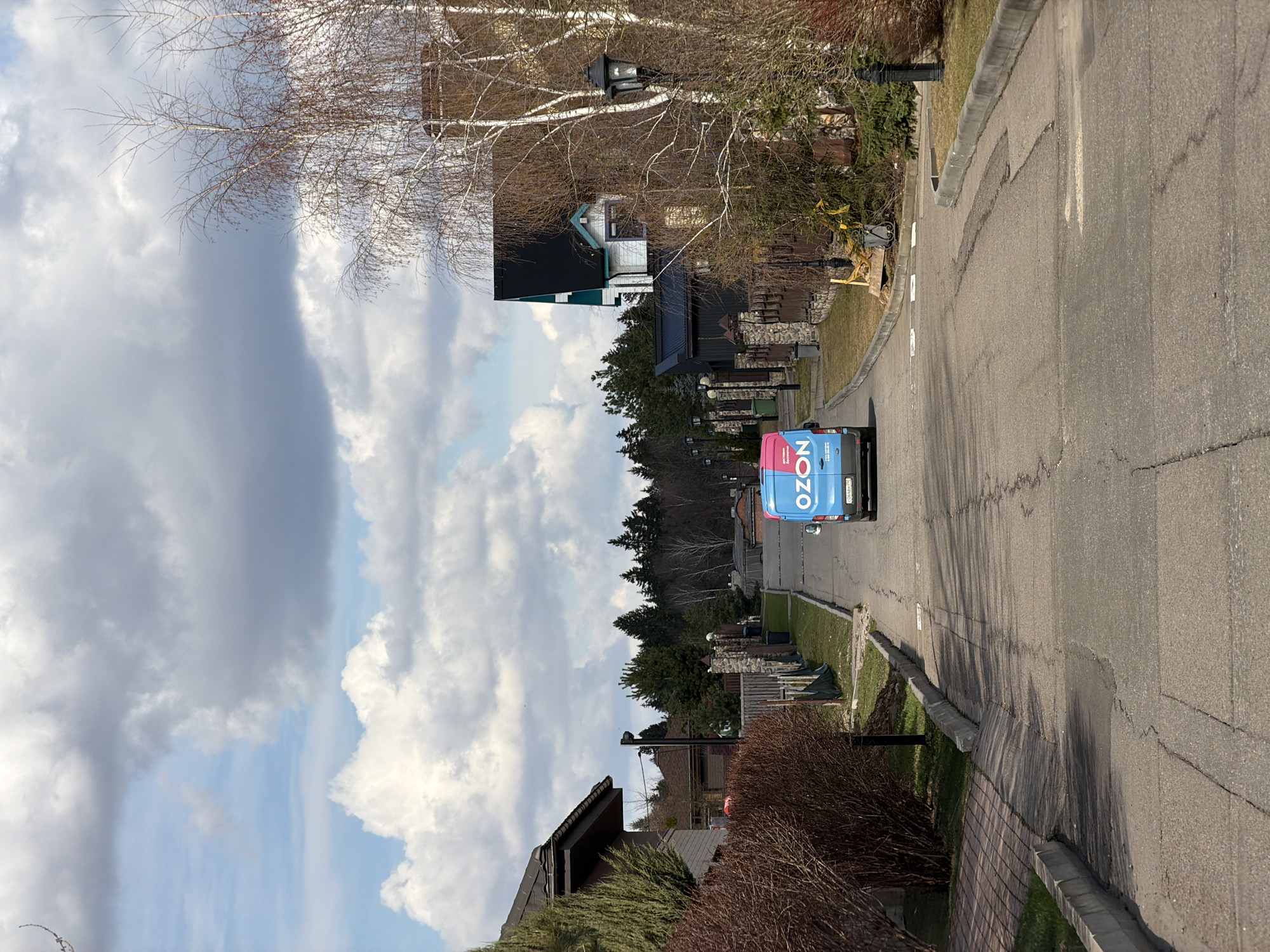A street scene in Mishutkino, Russia. An Ozön delivery van is driving on an asphalt road lined by sidewalks and houses. The sky is cloudy.
