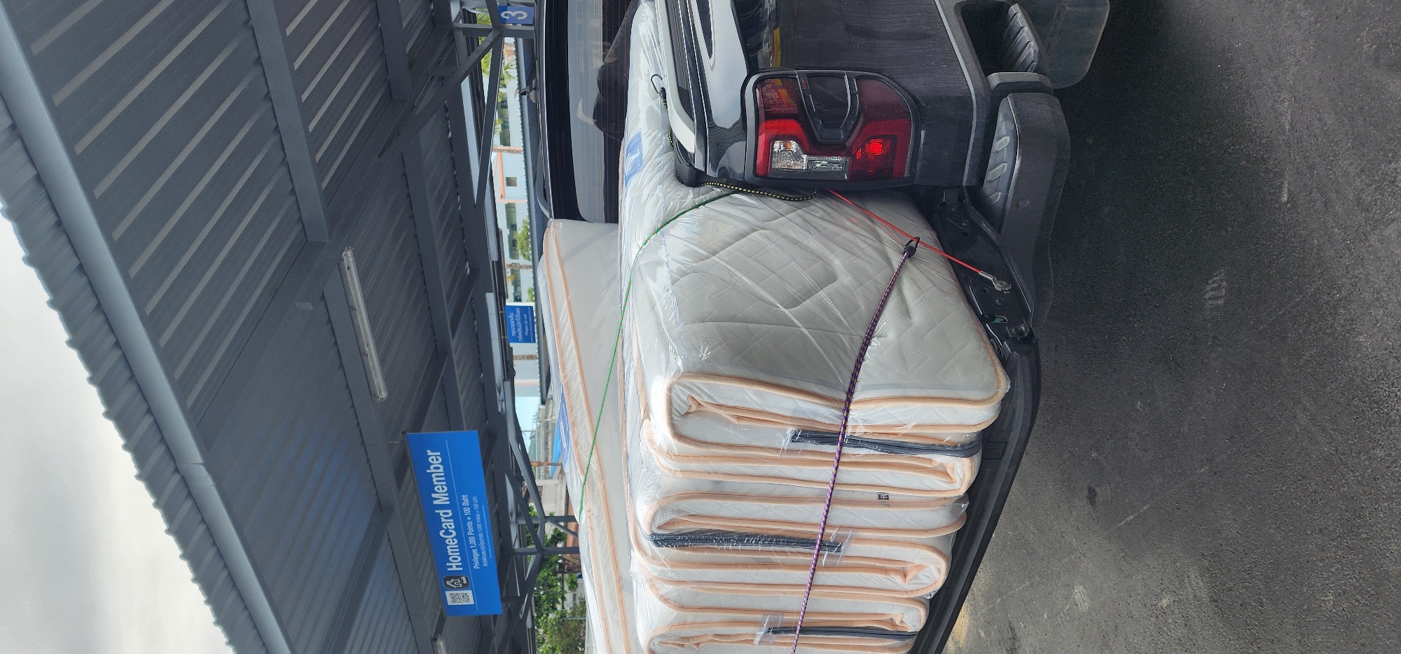 In Baan Chaweng Noi, Thailand, a black pickup truck is parked under a covered carport. The truck's bed is loaded with several tightly wrapped mattresses, secured with ropes and straps. In the background, a blue sign reads "HomeCard Member" and displays information about membership privileges. The scene appears to be a parking lot, indicated by the asphalt ground and overhead structure. The sky is overcast.