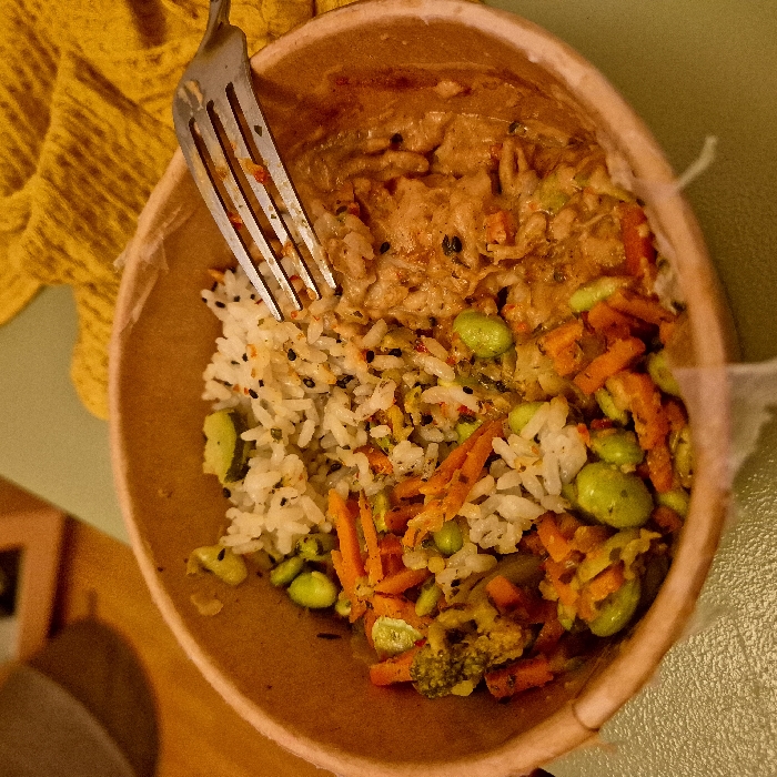 An overhead shot shows a tan bowl containing a meal. One side holds orange chicken with sauce. Adjacent is white rice speckled with black seeds, mixed with vibrant orange shredded carrots and green edamame. A fork pierces the left side of the rice. A yellow fabric sits beside the bowl. The bowl's surface has a rough, possibly biodegradable texture.