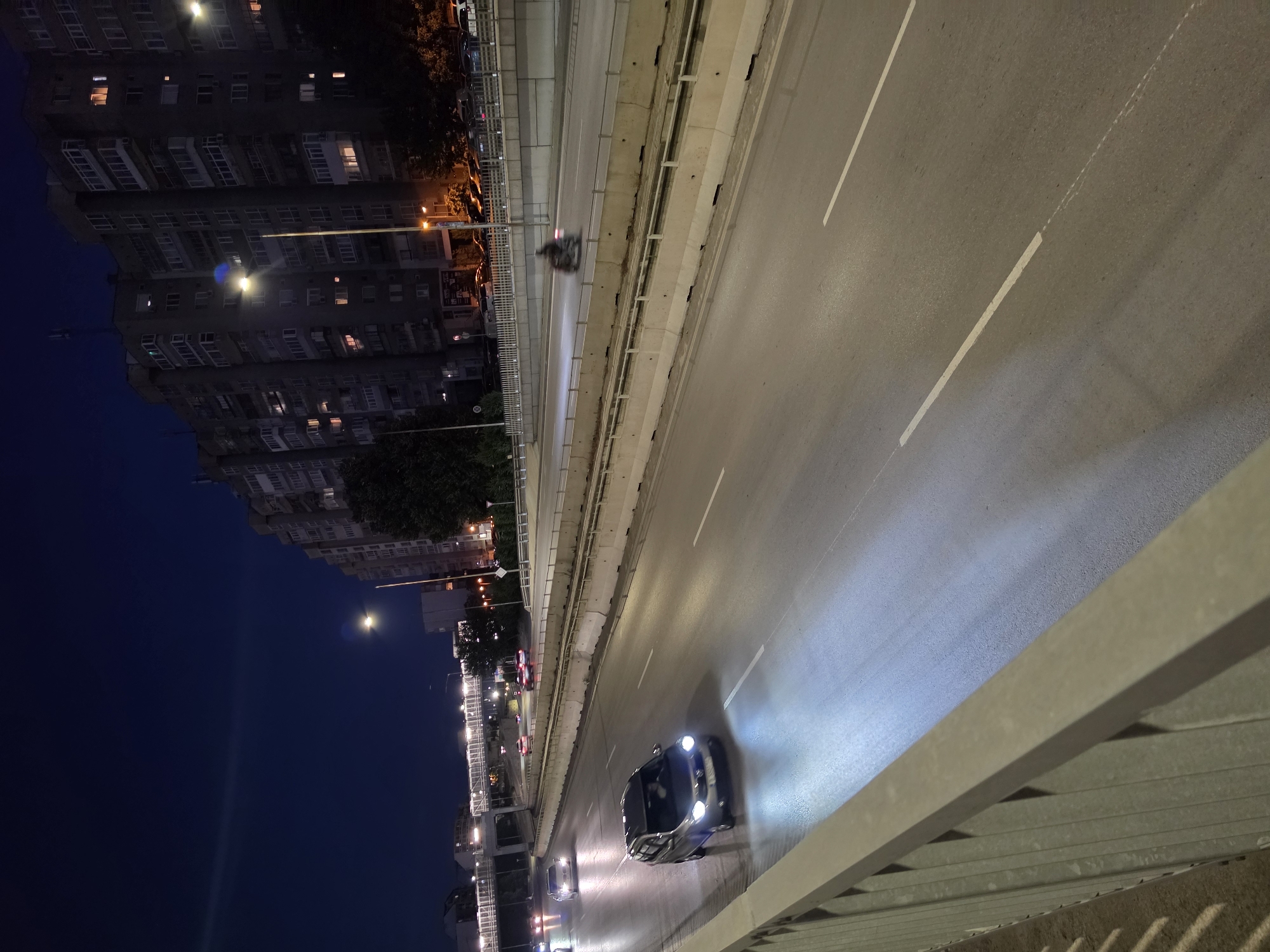 This image depicts a nighttime scene on a multi-lane highway likely within the city of Varna, Bulgaria. The foreground shows a concrete barrier with a metal railing, suggesting the vantage point is from an overpass or elevated walkway. The highway itself is illuminated by artificial lights, casting long reflections on the wet asphalt.
Several vehicles are visible on the highway. In the middle lane, a silver sedan with its headlights on is moving towards the viewer. Further down the highway, a motorcycle with a rider is visible in motion, appearing as a blurred silhouette due to its speed. Other vehicles are also present, some with their lights on, indicating ongoing traffic.
In the background, a large apartment building with numerous lit windows dominates the skyline, characteristic of urban residential areas. Trees and streetlights are also present, contributing to the city atmosphere. The sky is dark blue, indicating it is nighttime. The overall environment suggests a busy urban highway infrastructure.