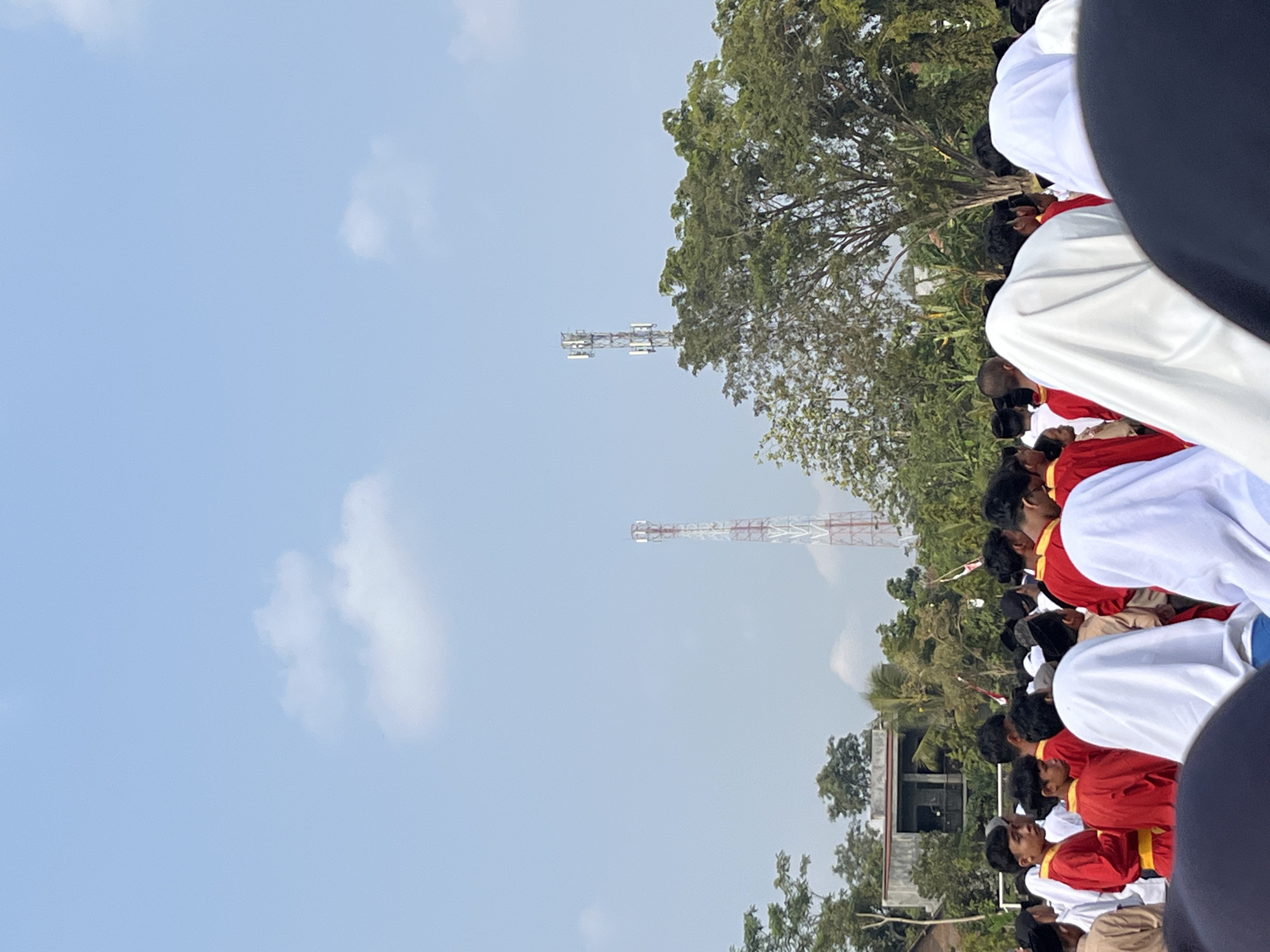 This image depicts an outdoor gathering of people, primarily young males, in Gembong, Indonesia. Many of the individuals are wearing red uniforms with yellow sashes, suggesting a ceremonial or organized event. Some are also wearing white head coverings or garments. In the background, two cellular towers are visible against a light blue sky with scattered clouds. Lush green trees and a building are also present in the background. The scene appears to be taking place during the daytime, with ample sunlight. The focus is on the group of people in the foreground, with the environment forming the backdrop.