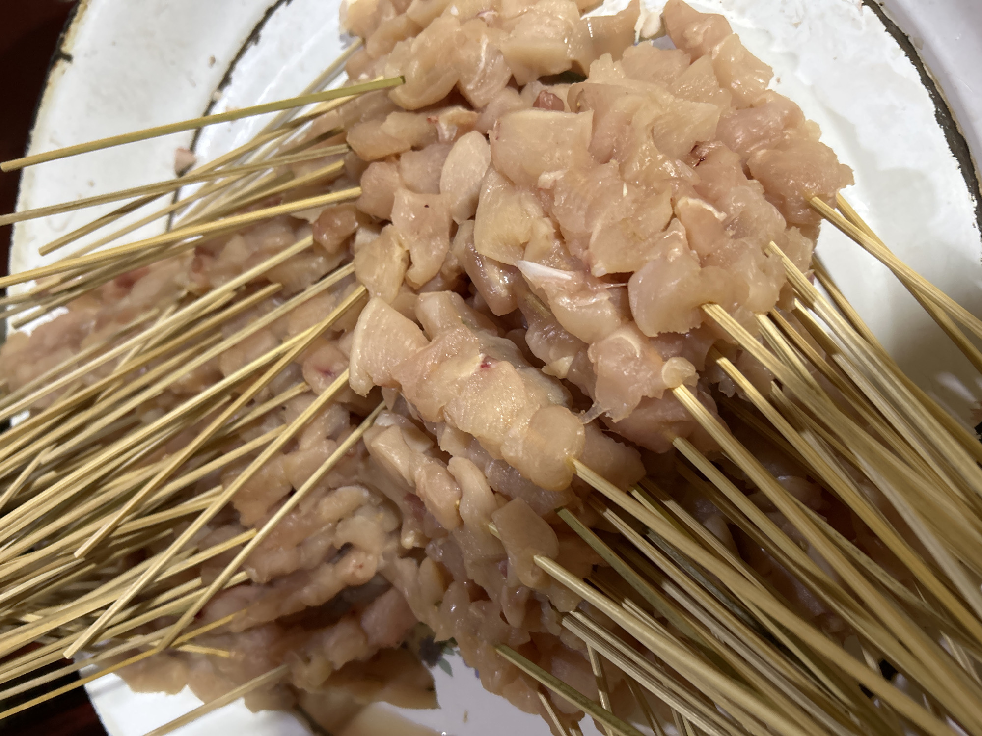 The image shows a close-up view of raw chicken skewers on a white plate. The chicken pieces are cut into bite-sized chunks and threaded onto thin bamboo skewers. The skewers are arranged in a dense cluster, with many of them overlapping. The chicken appears fresh and uncooked, with a pale pinkish-white color. The plate has a slightly worn, rustic appearance, with a dark rim that shows some signs of age or use. The background is slightly blurred, but appears to be a dark surface. The lighting is natural and diffused, highlighting the texture of the chicken and the grain of the bamboo skewers. There is no indication of any specific location in Depok, Indonesia, as the image focuses solely on the food preparation.