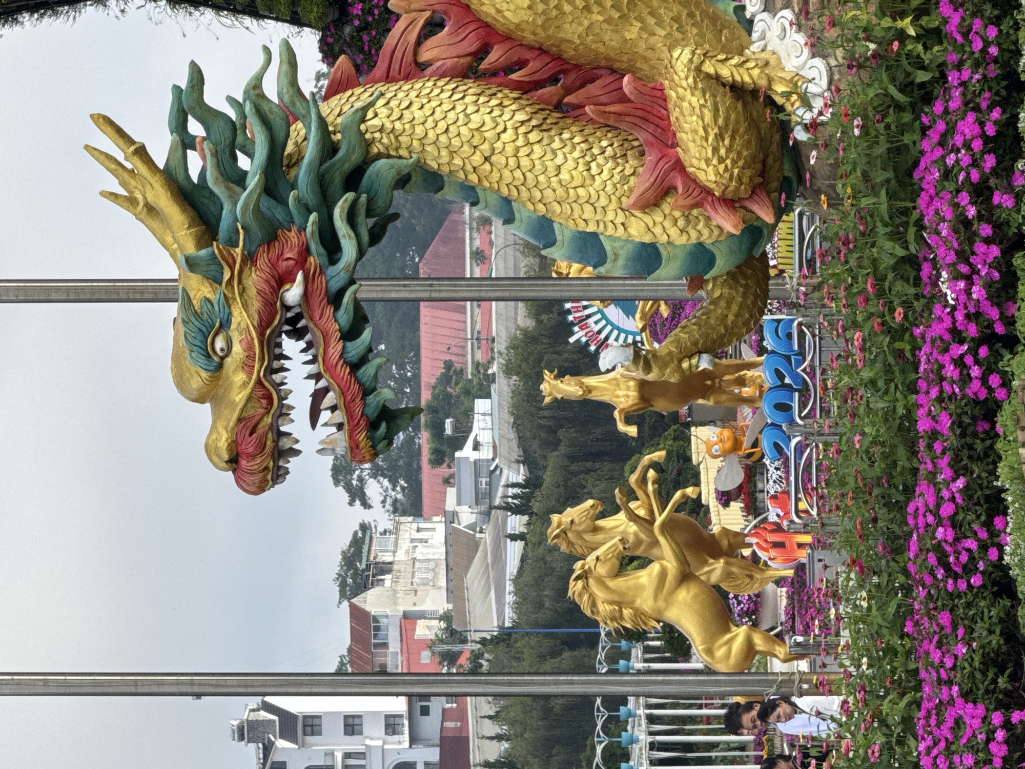 In Phường Xuân Hương - Đà Lạt, Vietnam, a large, vibrantly colored dragon statue dominates the foreground, its golden scales and green mane detailed. To the left of the dragon, three golden horse statues are positioned, two in rearing poses and one standing. In front of the horses and dragon, a floral arrangement of bright pink and purple flowers is displayed, with a sign that reads "2026" in blue lettering. Behind the statues, a collection of buildings with red-tiled roofs and white facades are visible, nestled amongst lush green trees. A large white building with multiple windows stands out on the left side of the frame. Several people are present, including two women looking towards the statues, and a man partially visible behind them. The sky is overcast.