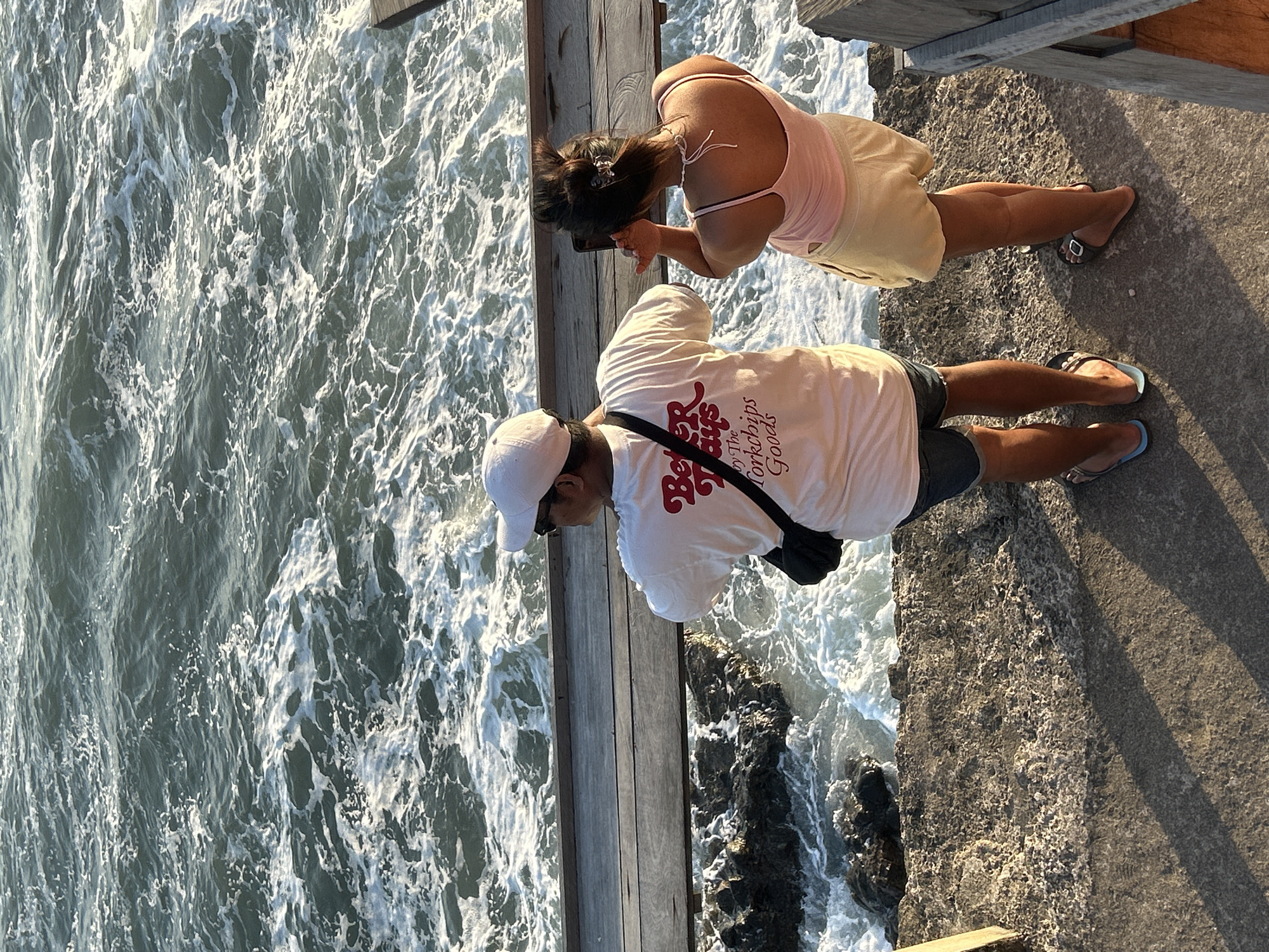 Two individuals are standing on a concrete ledge overlooking a churning ocean, with a wooden pier structure extending from the ledge and into the water. The person on the left, wearing a white cap and a white t-shirt with red text, is holding a phone or camera up to take a picture. The person on the right, wearing a pink tank top and light-colored shorts, is also holding a phone and appears to be taking a photo or video of the ocean. The water is a deep blue-green with significant white foam and waves crashing against dark rocks below the pier. The lighting suggests it is late afternoon or early morning, casting long shadows on the concrete ledge. The overall scene conveys a moment of enjoying the coastal scenery.