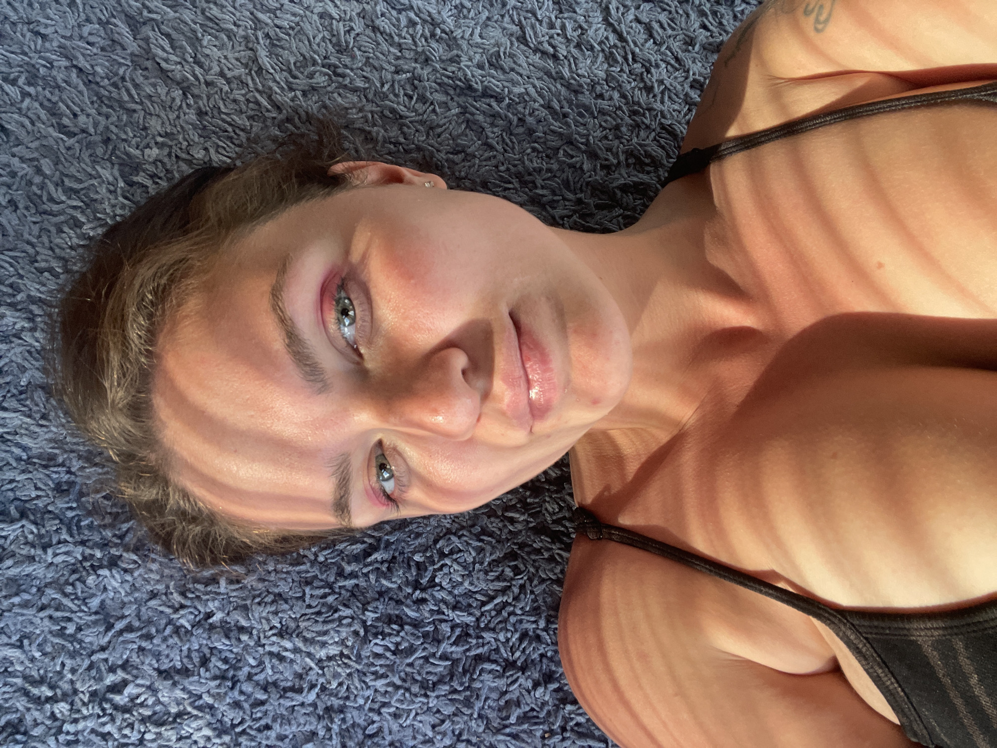 A woman lies on a blue carpet, angled slightly upward. She wears a black top. Light casts stripes across her face and body. Her eyes are visible, and she has light pink makeup on her eyelids. The setting appears to be indoors, possibly at Мосметаллоконструкция Онт, Russia.