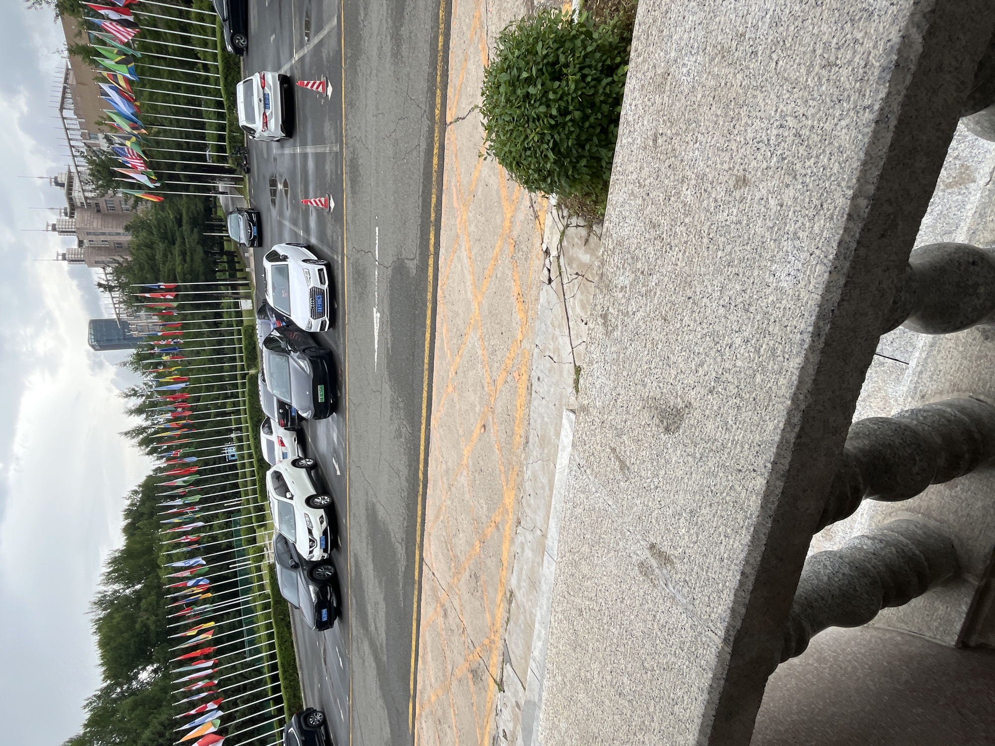 The image depicts a parking lot in front of a building adorned with a long row of international flags. Several cars are parked in designated spots, including a white Audi, a white SUV, a black Tesla, and other dark-colored sedans. The ground in the parking lot is asphalt with yellow painted markings. A concrete barrier with decorative stone pillars is in the foreground. In the background, trees line the area behind the flags, and modern buildings with numerous windows rise up against a cloudy sky. One of the buildings appears to be a research institution or university, with the flags suggesting an international gathering or diplomatic presence. The scene is consistent with the location of the Changchun Institute of Optics, Fine Mechanics and Physics (CIOMP) in Changchun City, China, known for hosting international conferences and events, often marked by displays of national flags.