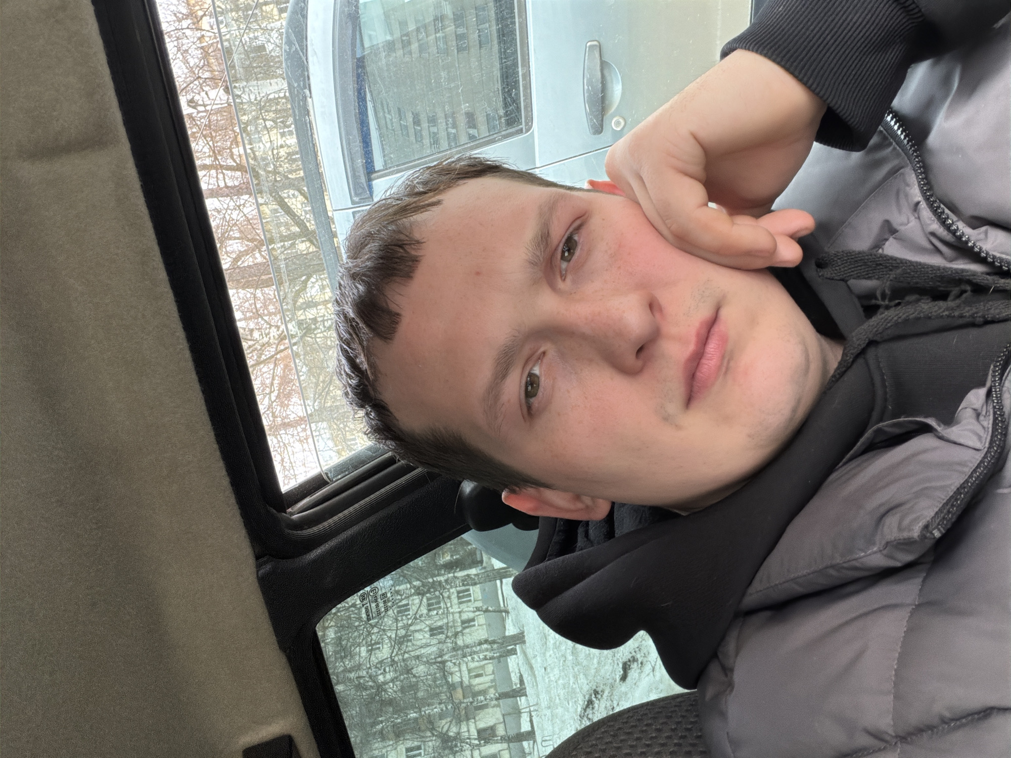 A photograph taken from inside a vehicle. A young adult male is the primary subject, positioned in the passenger seat. He is wearing a black hoodie under a gray puffer jacket and resting his hand on his face. His hair is short and brown. The view through the vehicle's windows shows a cityscape; barren trees and a pale gray building with multiple windows suggest a cold-weather environment. Outside the passenger side window, part of a silver vehicle is visible. Based on details visible through the window, it is probable this photo was taken in Izhevsk, Russia.