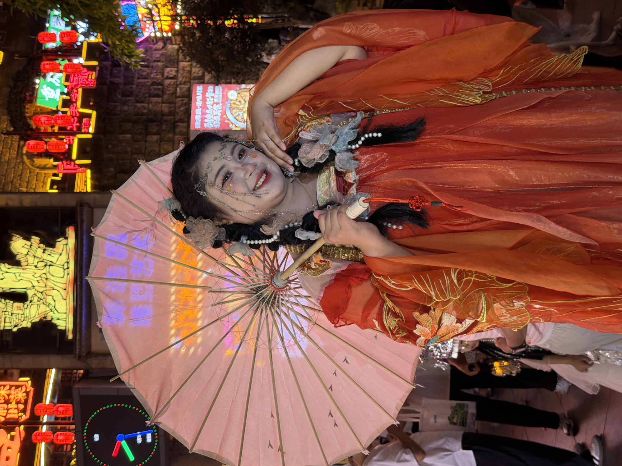 In Yuzhong District, China, a young woman stands in front of a brightly lit street scene at night. She is adorned in a vibrant orange and red traditional Chinese-style dress with gold embroidery. Her hair is styled in elaborate braids adorned with beads and flowers, and she wears ornate jewelry on her face and head. In her right hand, she holds a pale pink umbrella open above her. The background is bustling with activity and illuminated by numerous neon signs and red lanterns, indicative of a commercial or entertainment area. People are visible in the background, some holding bags or other items. The atmosphere suggests a festive or cultural event.