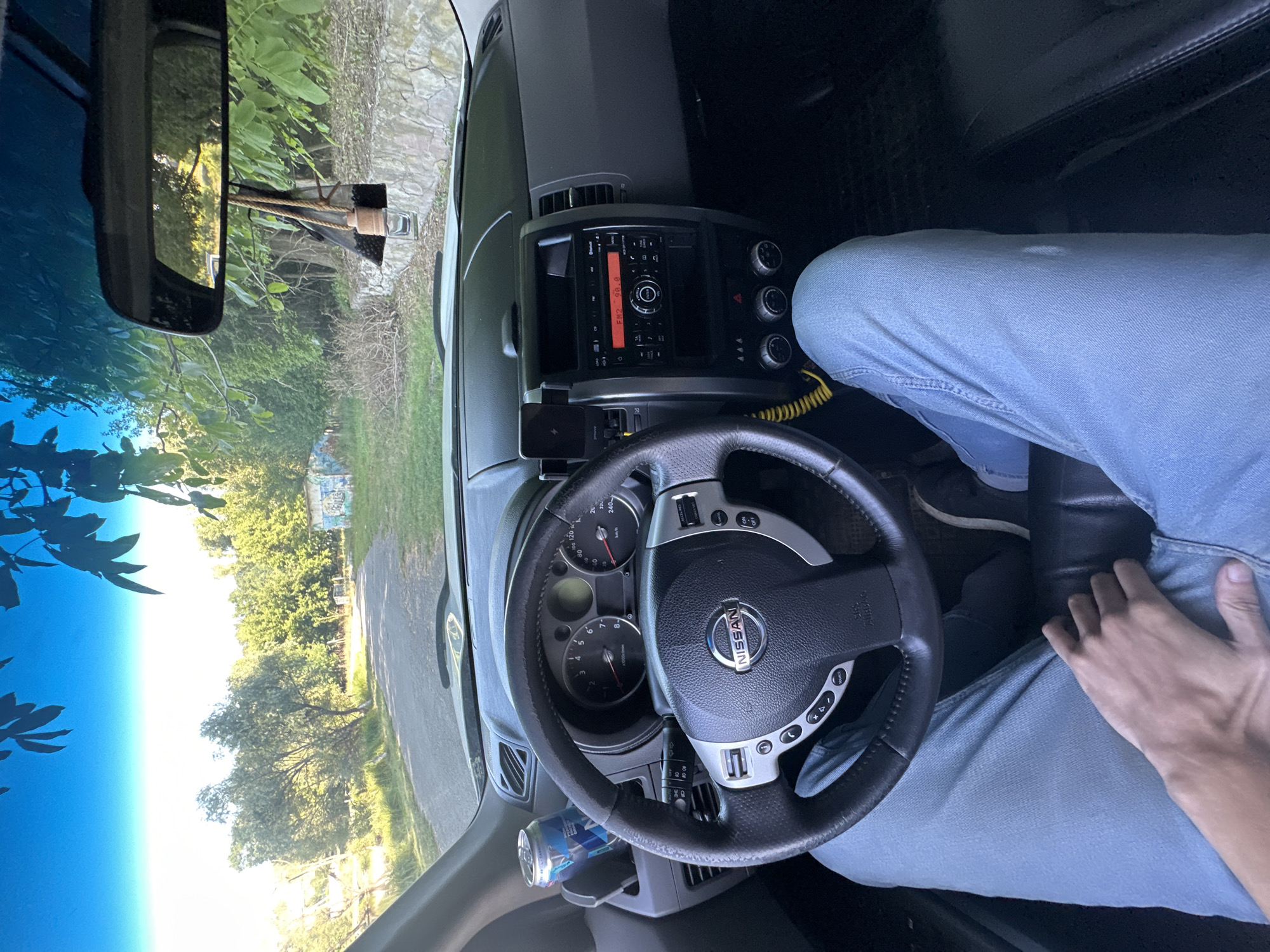 The image shows the interior of a Nissan X-Trail SUV, parked on a dirt road next to a grassy embankment lined with trees. The dashboard is visible, featuring a steering wheel with the Nissan logo, a radio, and climate controls. A can of drink is in a cup holder. Through the windshield, a rural landscape with trees and a cloudy sky is visible. The scene appears to be in or near the village of Makiv, Ukraine.