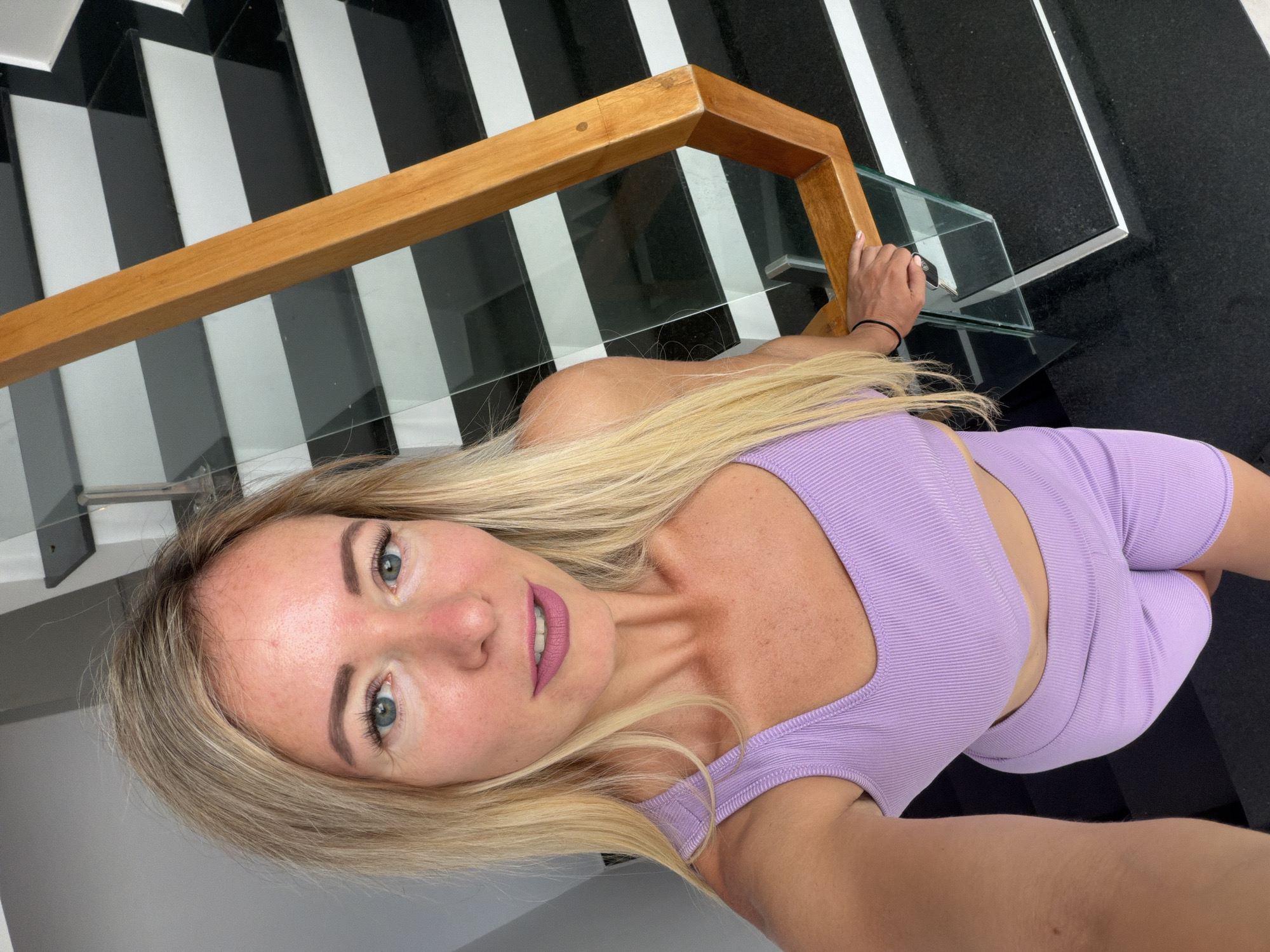 A blonde woman, wearing a lilac sports bra and shorts, takes a selfie on a modern staircase in what appears to be a residential building in Da Lat, Vietnam. The staircase has black and white striped steps with glass railings and a wooden handrail. The woman is looking directly at the camera, with her left hand resting on the handrail. The scene is brightly lit, suggesting daylight.