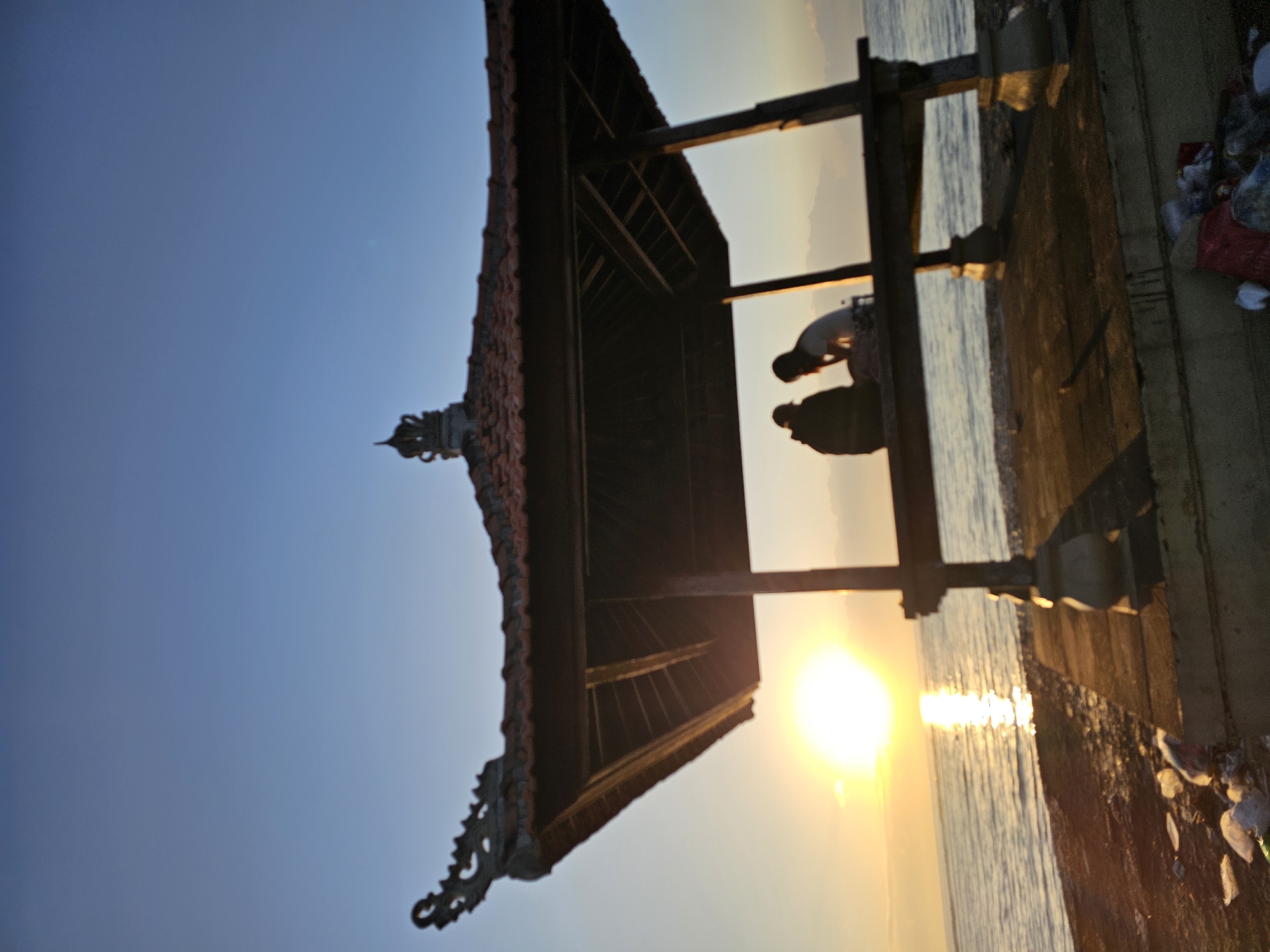 This is a photograph taken at sunrise on a beach in Denpasar, Indonesia. In the foreground, there is a wooden gazebo with a tiled roof, featuring intricate carvings on the eaves. Two individuals are silhouetted within the gazebo, sitting and facing the ocean. One person appears to be wearing a dark hooded garment, while the other is wearing a lighter shirt. The sun is rising over the ocean, casting a bright, warm glow and reflecting on the water. The beach area in front of the gazebo is littered with debris, including plastic bags and other trash. The ocean is calm, with gentle waves lapping the shore. The sky is a clear blue, transitioning to a lighter hue near the horizon where the sun is setting.