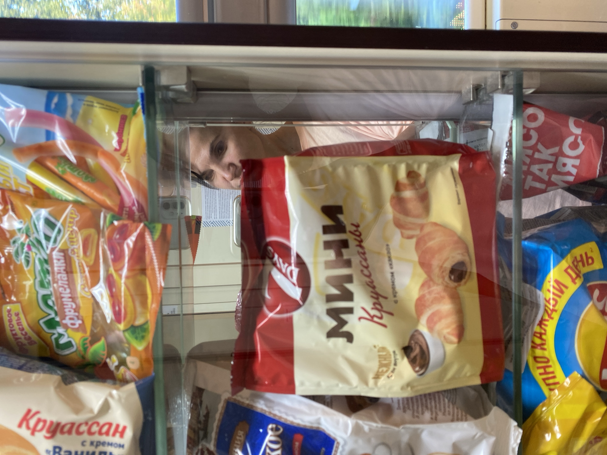 A woman is visible through the glass door of a refrigerated display case, which contains various food items. The display case appears to be a commercial establishment, likely a store or a vending machine, judging by the arrangement of packaged goods. The woman, with dark hair and wearing a light pink t-shirt, is looking to her left, with her face partially obscured by the display.

The products in the display case include bagged pastries, such as croissants, with Russian text visible on the packaging. One prominent package features the word "МИНИ" (MINI) and "Круассаны" (Croissants) in large, stylized lettering, along with an image of croissants and a bowl of chocolate cream. Another bag on the left has text such as "Круассан" and "Ваниль" (Vanilla). Below the display, other bagged snacks are visible, one with a bright blue and yellow design and Russian text that includes "ПО КАЖДЫЙ ДЕНЬ" (every day). Another red bag with white text "ТАК МЯСО" (so meat) is also visible. The background, seen through the glass, suggests an indoor environment with some shelving and possibly other parts of the establishment. The lighting is artificial, illuminating the products within the case.

The context provided specifies the location as Gryazi, Russia. This suggests the establishment is a retail outlet in that city, potentially a convenience store, supermarket, or a food service area within a larger facility. The overall scene depicts a moment of observation or selection of food items by a customer in a retail setting.