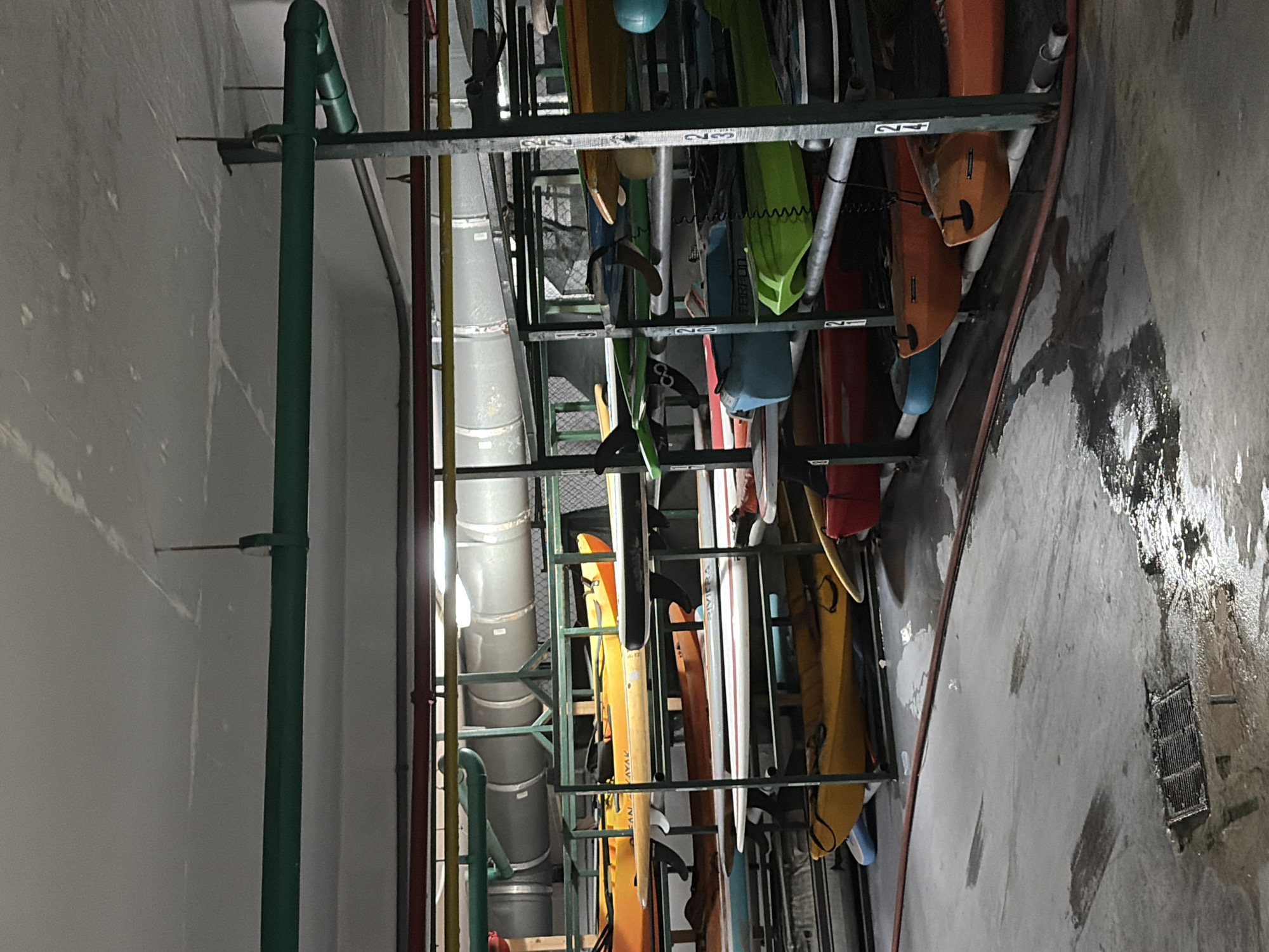 The image shows a storage area for kayaks and surfboards in a building. There is a metal rack system holding the watercraft, which are of various colors and sizes. A large, segmented ventilation duct runs parallel to the rack. The location appears to be an indoor storage facility. The presence of water on the floor and a floor drain suggests this area may be prone to moisture or drainage issues. There is no indication of people or interaction in the image. No evidence directly connects the scene to Key Biscayne, United States of America.