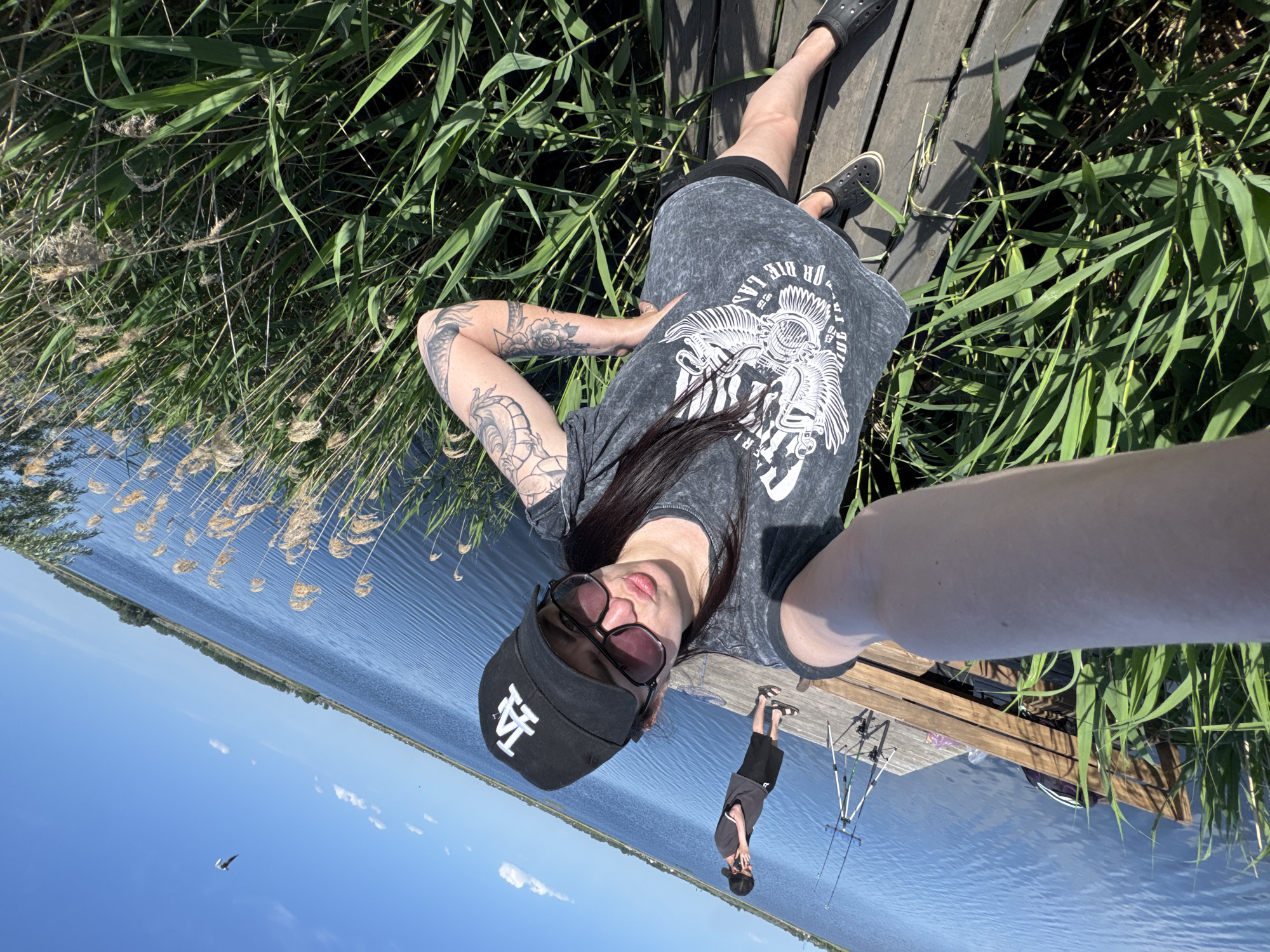 A woman lies on a wooden dock, wearing a graphic t-shirt, black shorts, black shoes, a black baseball cap with "LA" on it, and sunglasses. Her arms are tattooed. She is holding the camera and taking a selfie. Beside the dock is tall grass and a large body of water reflecting the blue sky. There is a small figure of a man holding a fishing rod and what appears to be fishing poles nearby, reflecting in the water. A bird is visible in the blue sky. Location is not specified in the image.