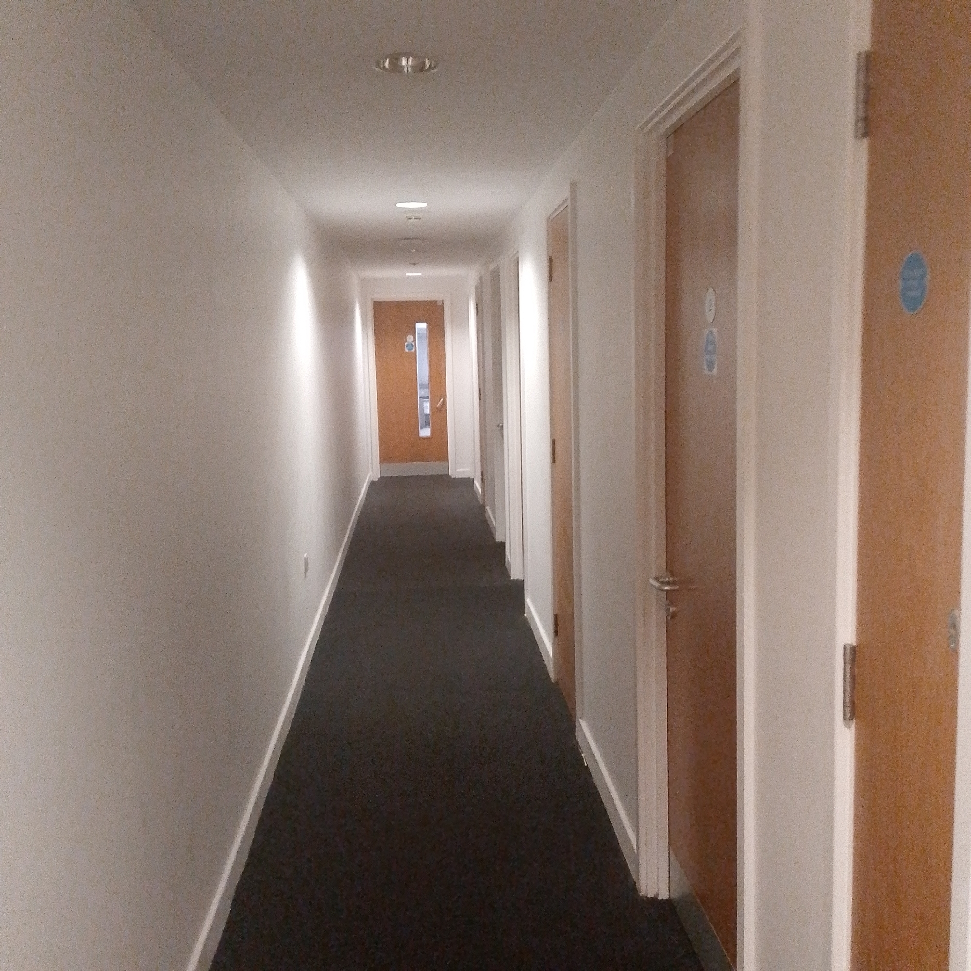 The image shows a long, narrow hallway in a building in Coventry, United Kingdom. The walls are painted white, and the floor is covered with dark grey carpet. There are several closed wooden doors on the right side of the hallway, some with signs on them. The hallway is lit by ceiling lights. The view is looking down the hallway towards a distant door. There are no people visible in the image.