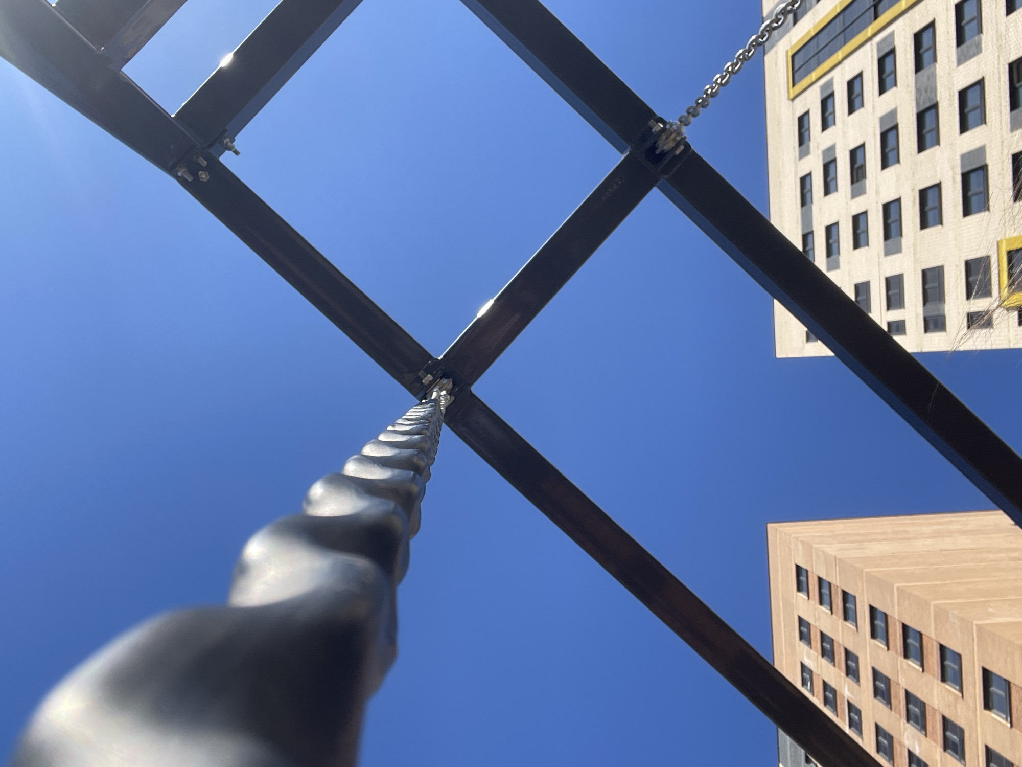 The image presents a low-angle view of a metal structure with a chain, set against a clear blue sky. Two buildings with multiple windows are visible in the background. The metal structure appears to be a part of a climbing frame or similar apparatus. The specific location in Izhevsk, Russia, is not identifiable from the image alone. There are no people visible. The scene consists of static objects within an outdoor environment.