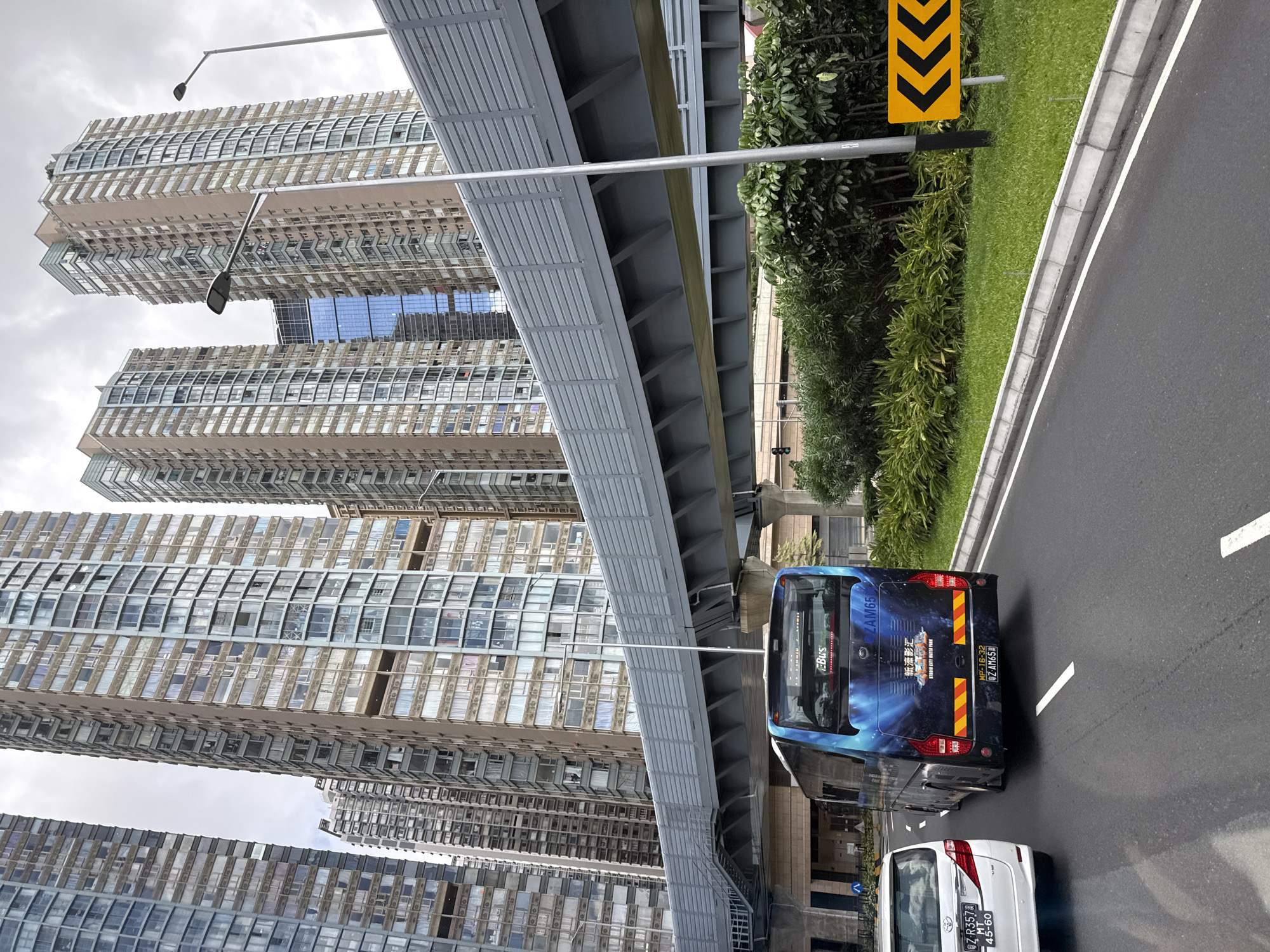 This is a photograph taken in Macau, China, featuring a view of tall residential buildings and a multi-level overpass system. The foreground shows a road with a white van on the left and a blue bus with a vibrant graphic on the back driving on the right. The bus displays the license plate "Z AM65" and has text indicating "STORM CITY WATER PARK." The background is dominated by numerous high-rise apartment buildings with numerous windows, suggesting a densely populated urban area. A gray, elevated walkway or barrier runs across the middle of the image, supported by concrete pillars. Lush green vegetation lines the side of the road. The sky is overcast.