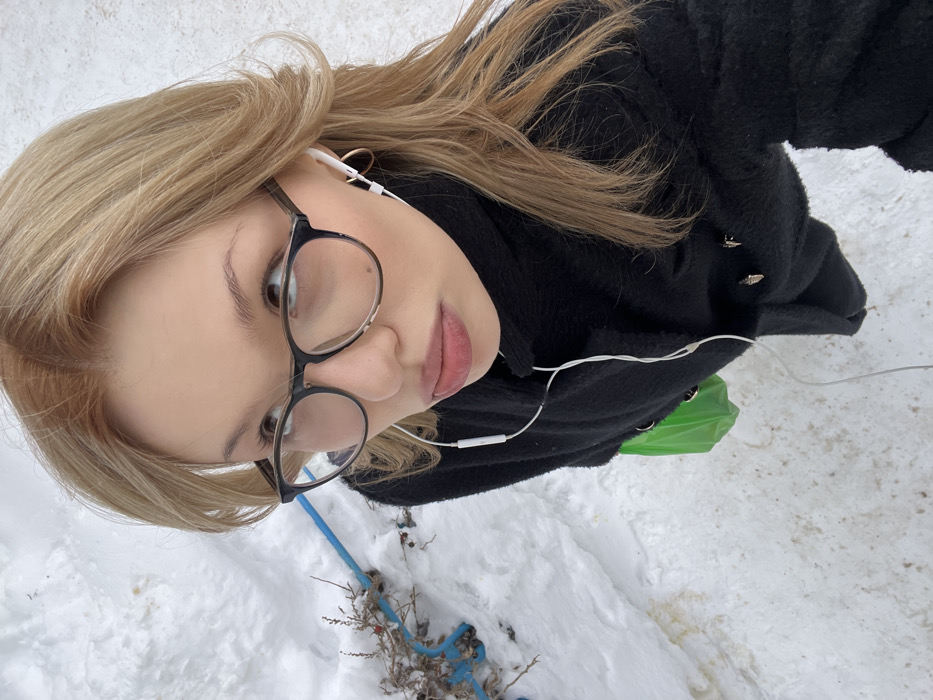 This image captures a female individual outdoors in a snowy environment, consistent with winter conditions in the city of Chaikovsky, Russia. The subject is depicted from a high-angle perspective, facing generally upwards and to her right. She has blonde hair with darker roots, wears black-framed eyeglasses, and has white wired earphones in both ears, with the wire extending downwards. A small gold hoop earring is visible in her right ear. Her lips are full and slightly parted. She is wearing a black, textured winter coat with a high collar and visible silver-colored fastenings or embellishments. A bright green plastic bag is visible on her left side, partially obscured by the coat. The ground is covered in white snow, featuring patches of exposed dirt or melted snow. To the left of the subject, a bare shrub with small red elements (possibly berries or dried leaves) protrudes from the snow. A bright blue flexible pipe is partially visible in the snow, extending horizontally near the shrub.