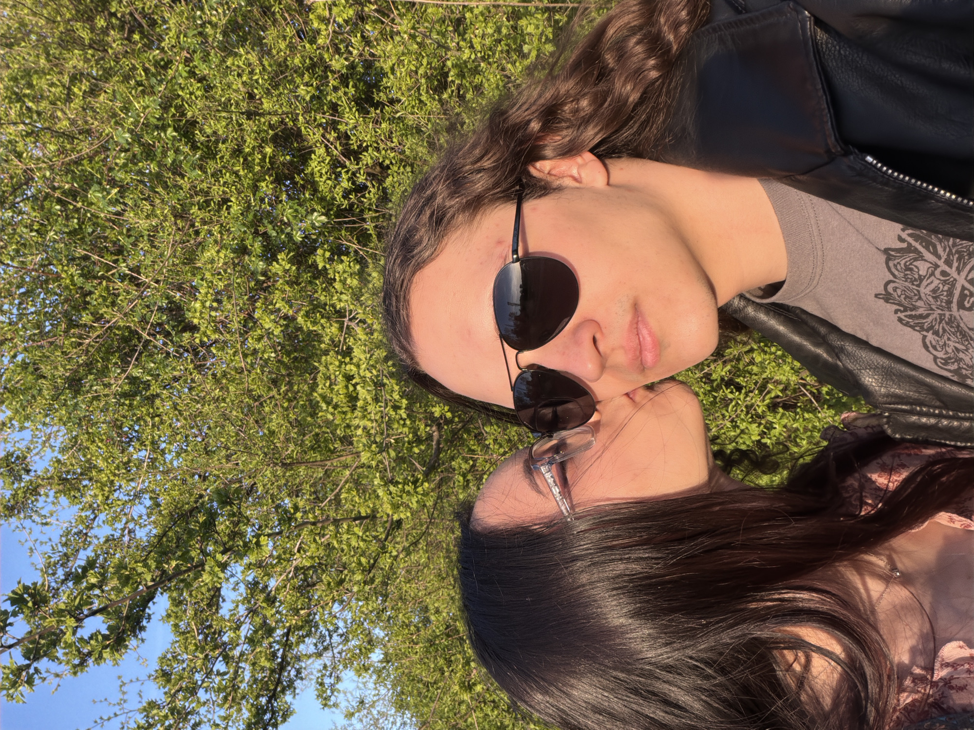 Two people are kissing. The person on the top is wearing sunglasses, a black leather jacket, and a grey shirt. They have long brown curly hair. The person on the bottom has long dark hair and is wearing glasses and a patterned shirt. They are outside, in front of a tree with green leaves and a blue sky. It is not possible to determine the location in the city of Sofia based on the image.