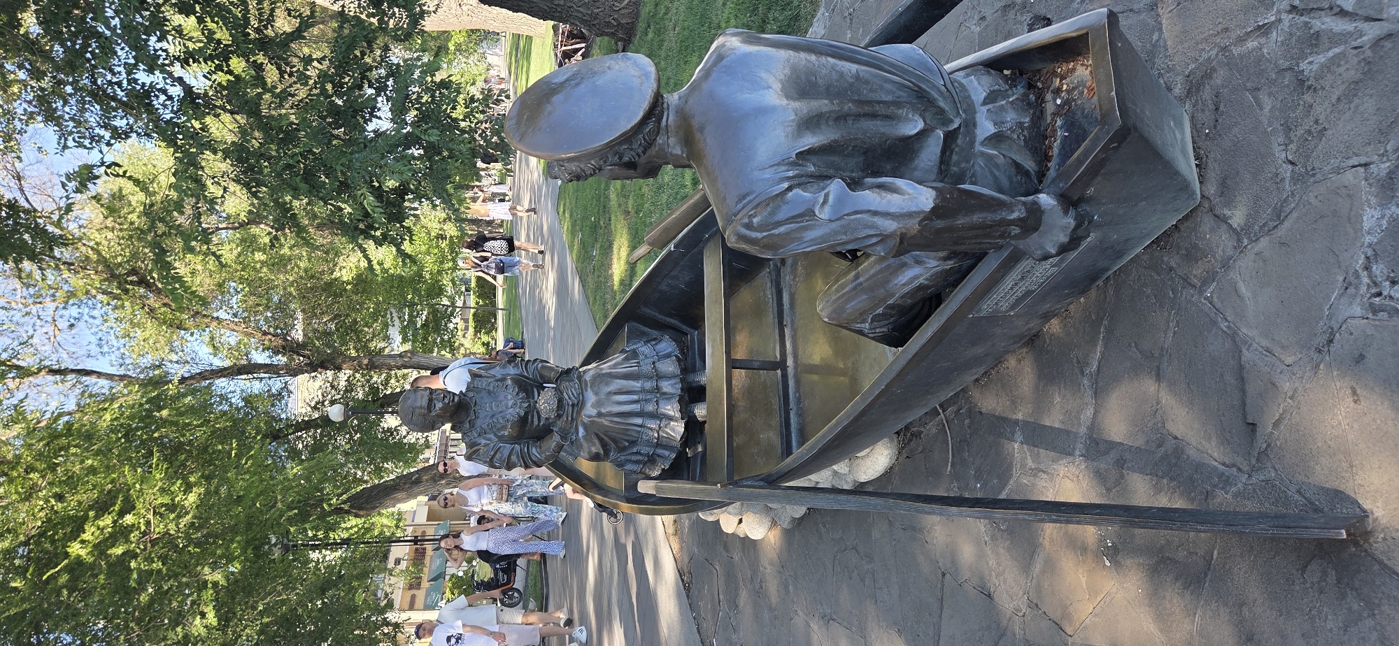 The image depicts a bronze sculpture of a man and a woman in a boat, situated in a park in Rostov-on-Don, Russia. The sculpture is positioned on a paved walkway with a stone surface. The man, facing away from the viewer, is dressed in a uniform with a cap and appears to be rowing the boat. The woman, facing the viewer, is seated and dressed in period clothing, holding a bouquet of flowers. In the background, trees provide shade, and several people are walking along the path. The lighting suggests it is daytime, likely with sunlight filtering through the leaves.