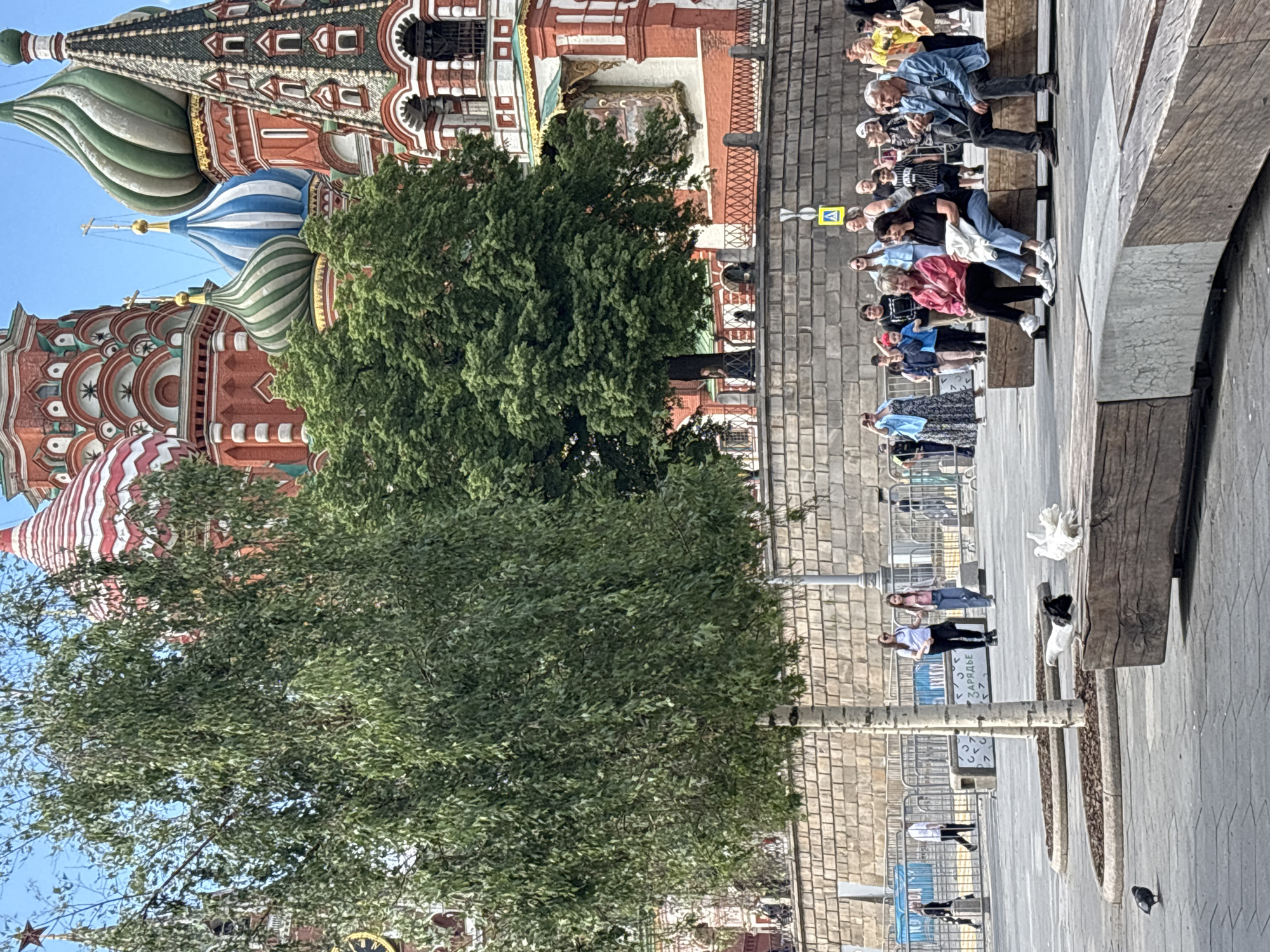 The image depicts a view of St. Basil's Cathedral in Moscow, Russia. Parts of the building's colorful domes and facades are visible behind dense green trees. A crowd of people is standing along a walkway adjacent to a brick wall. Some are seated on a wooden bench. The walkway also features trees, metal fences, light poles, and pigeons.