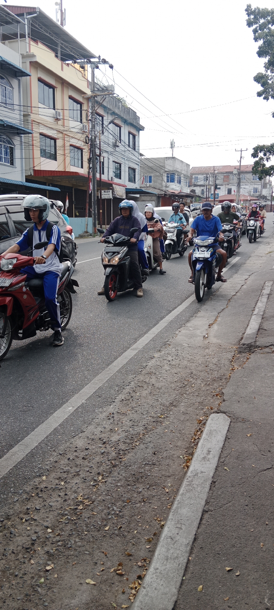 The image shows a street scene in Tanjung Pinang, Indonesia, with several motorbikes lined up on the road. The motorbikes are of various colors and models, and some of them are carrying passengers. The people on the motorbikes are wearing helmets, and some of them are wearing casual clothing. The road is paved with asphalt, and there are some trees and buildings on the side of the road. The sky is cloudy, and the overall lighting is overcast. The scene appears to be a typical day in a bustling Indonesian city, with motorbikes being a common mode of transportation. The architecture of the buildings suggests an urban environment, possibly a commercial area. The presence of power lines and utility poles is also characteristic of city infrastructure. The ground is littered with dried leaves, indicating a potential season or simply natural debris accumulation.