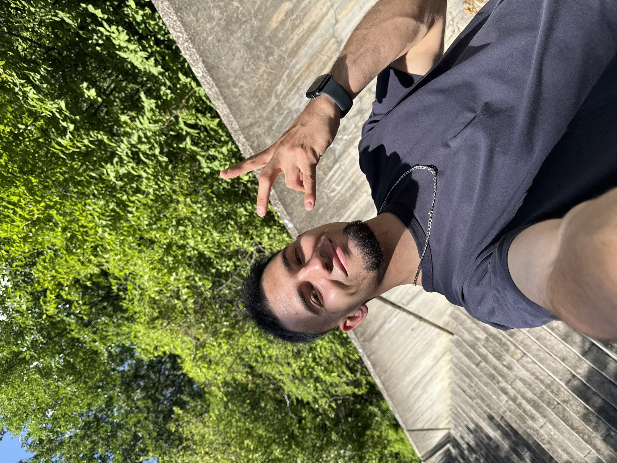 A young man with short dark hair and a goatee is taking a selfie outdoors. He is wearing a dark t-shirt and a silver chain necklace. His right hand is raised with two fingers extended in a rock and roll gesture. He is wearing a dark smartwatch on his left wrist. The background consists of lush green trees and a concrete structure that appears to be part of a staircase or amphitheater. The lighting suggests it is daytime. The location is the park area near the Monument to the Bulgarian-Soviet Friendship in Shumen, Bulgaria.