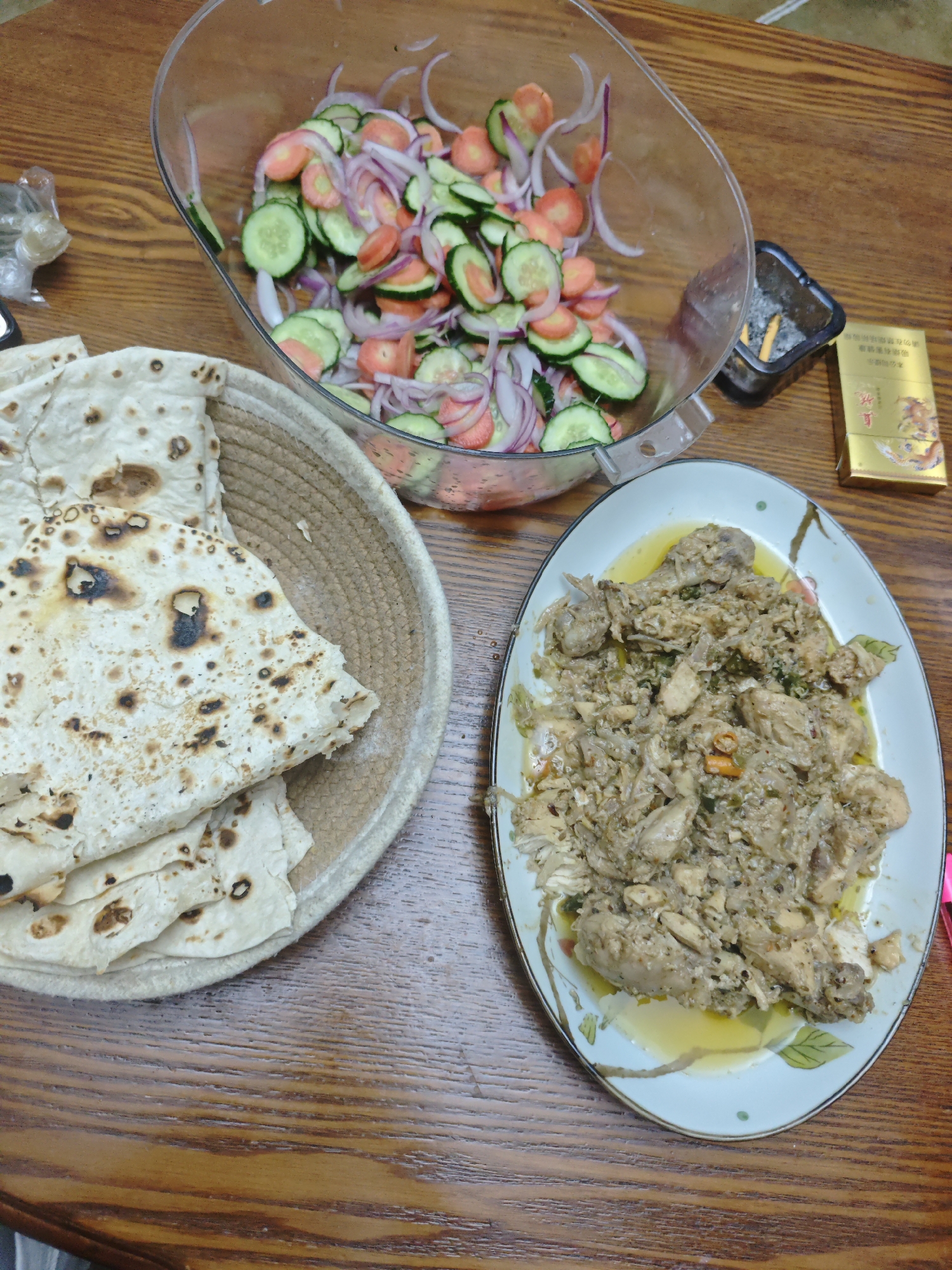 On a wooden table, a meal is laid out consisting of flatbread in a woven basket, a salad of sliced cucumbers, carrots, and red onions in a clear bowl, and a plate of what appears to be chicken. An ashtray containing cigarette butts and a pack of cigarettes are also on the table. The location is indicated as Chongqing High-tech Zone, China.