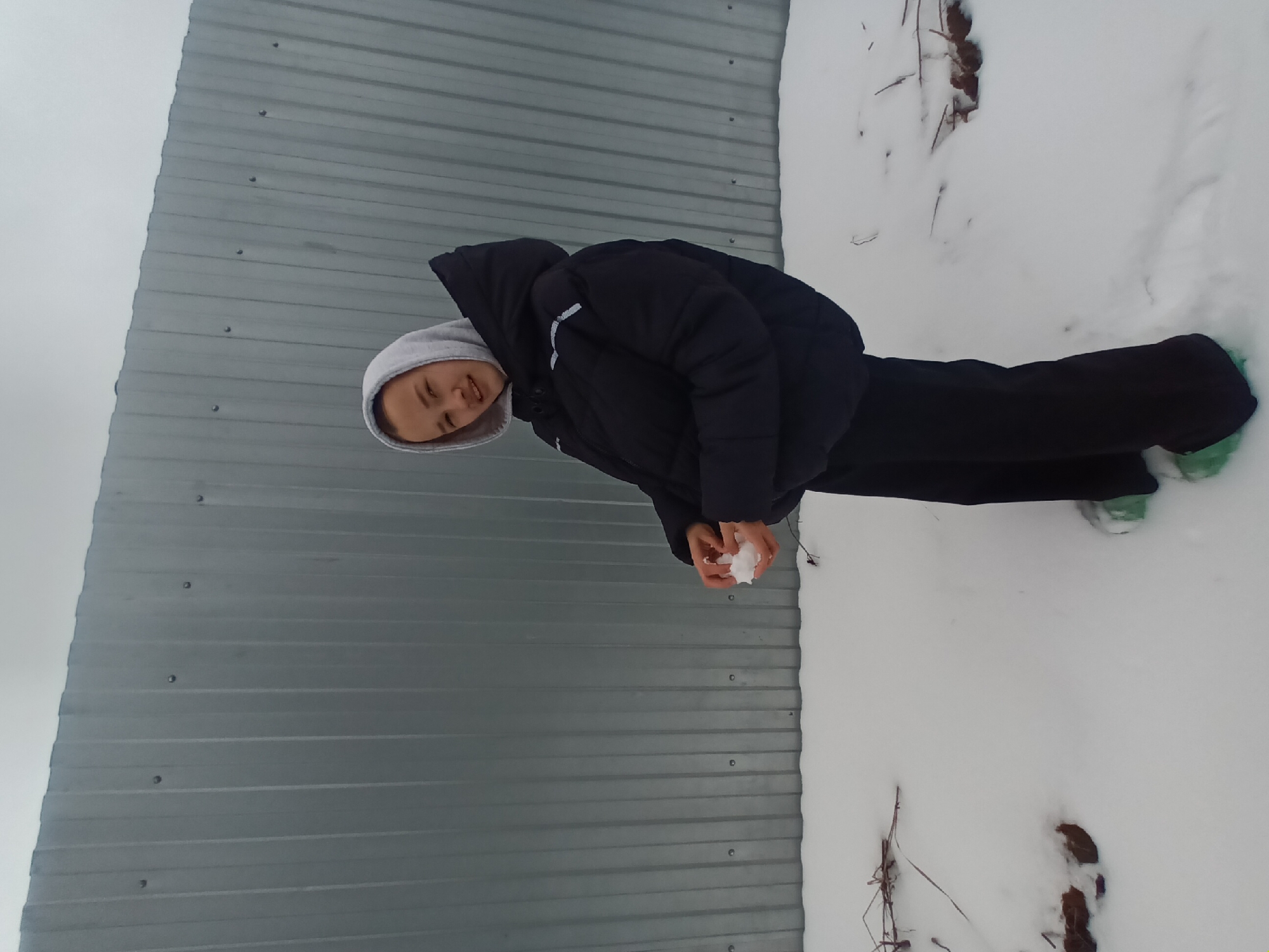 A person wearing a grey hooded sweatshirt underneath a black puffy jacket and dark pants stands in the snow near a grey corrugated metal fence. The person is holding a snowball in their hands. The scene appears to be in a snowy outdoor environment in the city of Ольховка, Russia.