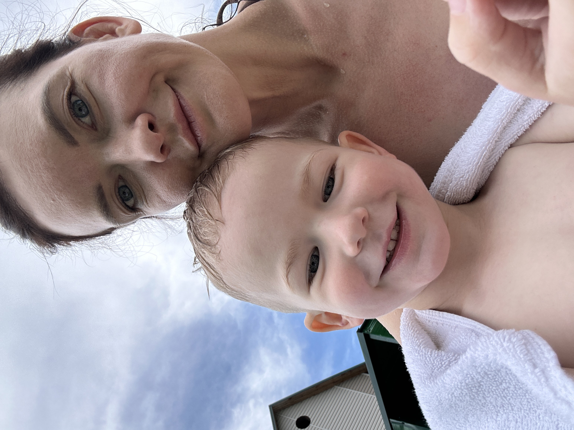 A selfie taken outdoors shows a woman with blue eyes and a woman with light brown hair smiling. A young child with light brown hair is in the foreground, a white towel draped around his shoulders. In the background, a building with beige siding and a circular window is visible. The sky is cloudy with patches of blue. The location is the city of Даниловка (алтайский), Russia.