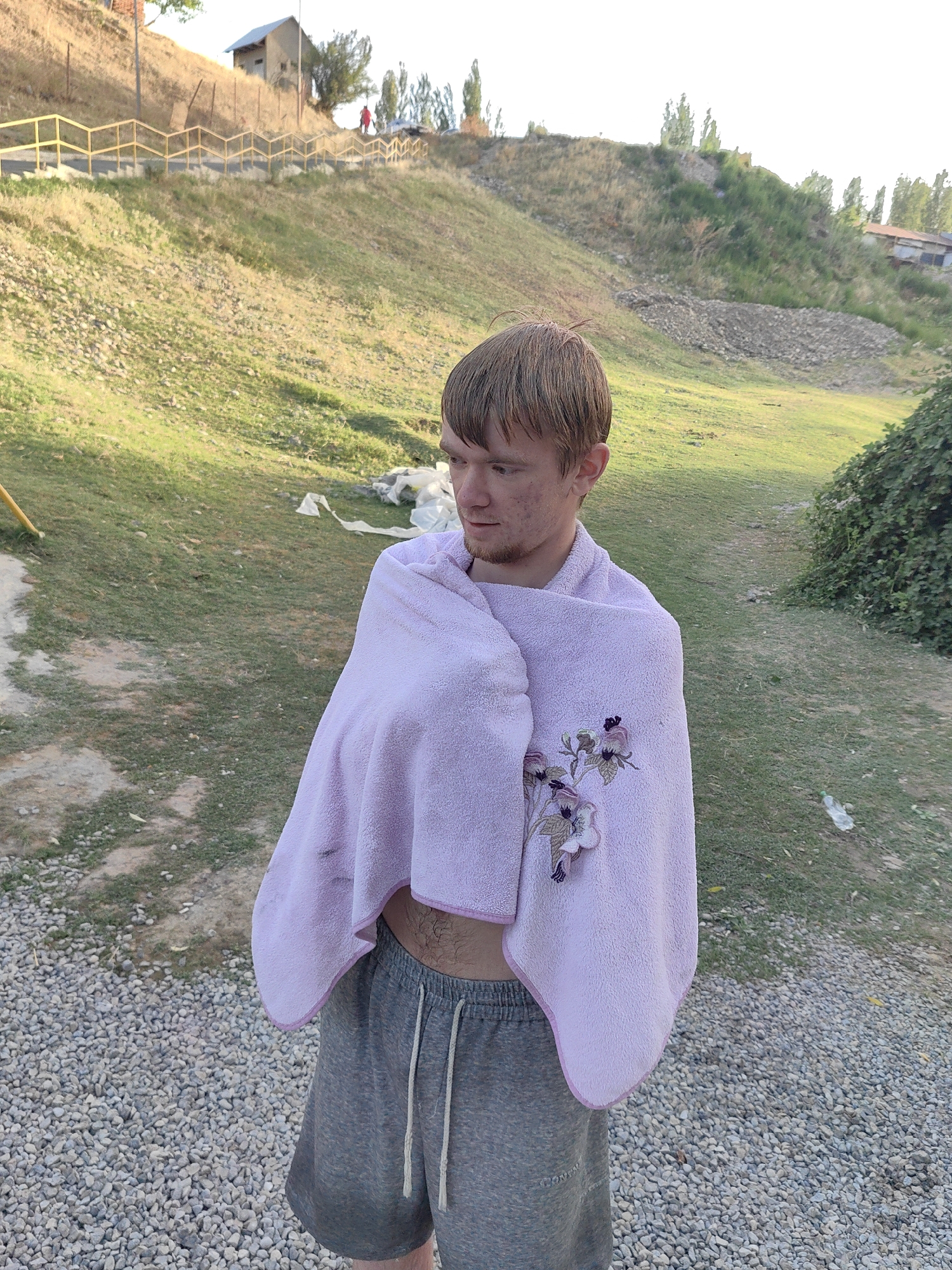 A young Caucasian male with light brown hair, light skin, and light stubble is wrapped in a light purple towel adorned with embroidered purple flowers. He is wearing gray athletic shorts. He stands in what appears to be an outdoor area with gravel on the ground in the foreground and a grassy slope behind him. In the background, a hill rises, with a small building and a yellow railing along a path visible near the top. Trees and more buildings are scattered on the distant hills. The overall setting suggests a rural or semi-rural environment.