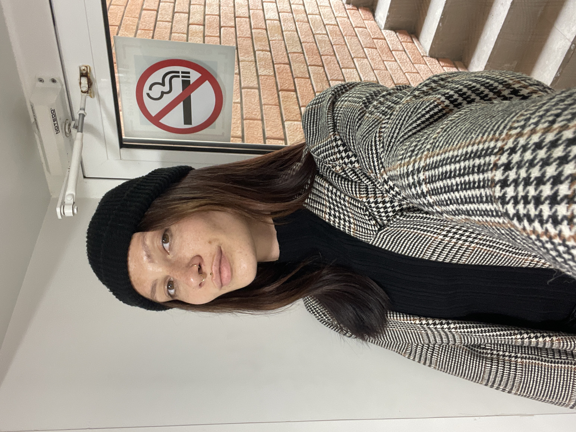 A woman is standing near a window with a "no smoking" sign. The background shows a brick wall and concrete stairs. The woman is wearing a black beanie, a black top, and a patterned houndstooth jacket. She has a nose ring. The window hardware is marked "NOTEGOO". The location appears to be Izhevsk, Russia.
