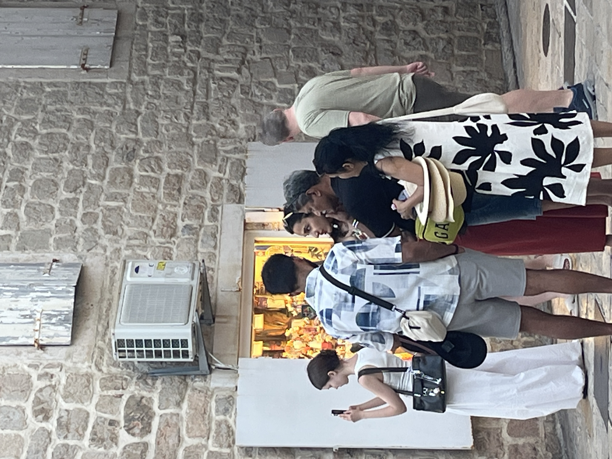A group of people are gathered outside a shop in Budva, Montenegro. The shop has a brightly lit interior displaying various items, and a large white shutter is positioned to the left of the shop entrance. An air conditioning unit is mounted on the stone wall above the shutter.
In the foreground, a young woman in a white dress is holding a smartphone and taking a picture. To her right stands a man wearing a blue and white patterned shirt and grey shorts. He has a dark bag slung across his shoulder. Next to him, a woman in a red dress appears to be talking to a man in a black shirt. Another woman, wearing a white dress with a black floral pattern, stands further to the right, holding a stack of straw hats. Behind her, a man in a green t-shirt and shorts is walking away from the viewer.
The building's exterior is constructed from rough-hewn stone blocks. The ground is paved with light-colored stones. A closed wooden shutter is visible on the upper part of the building.