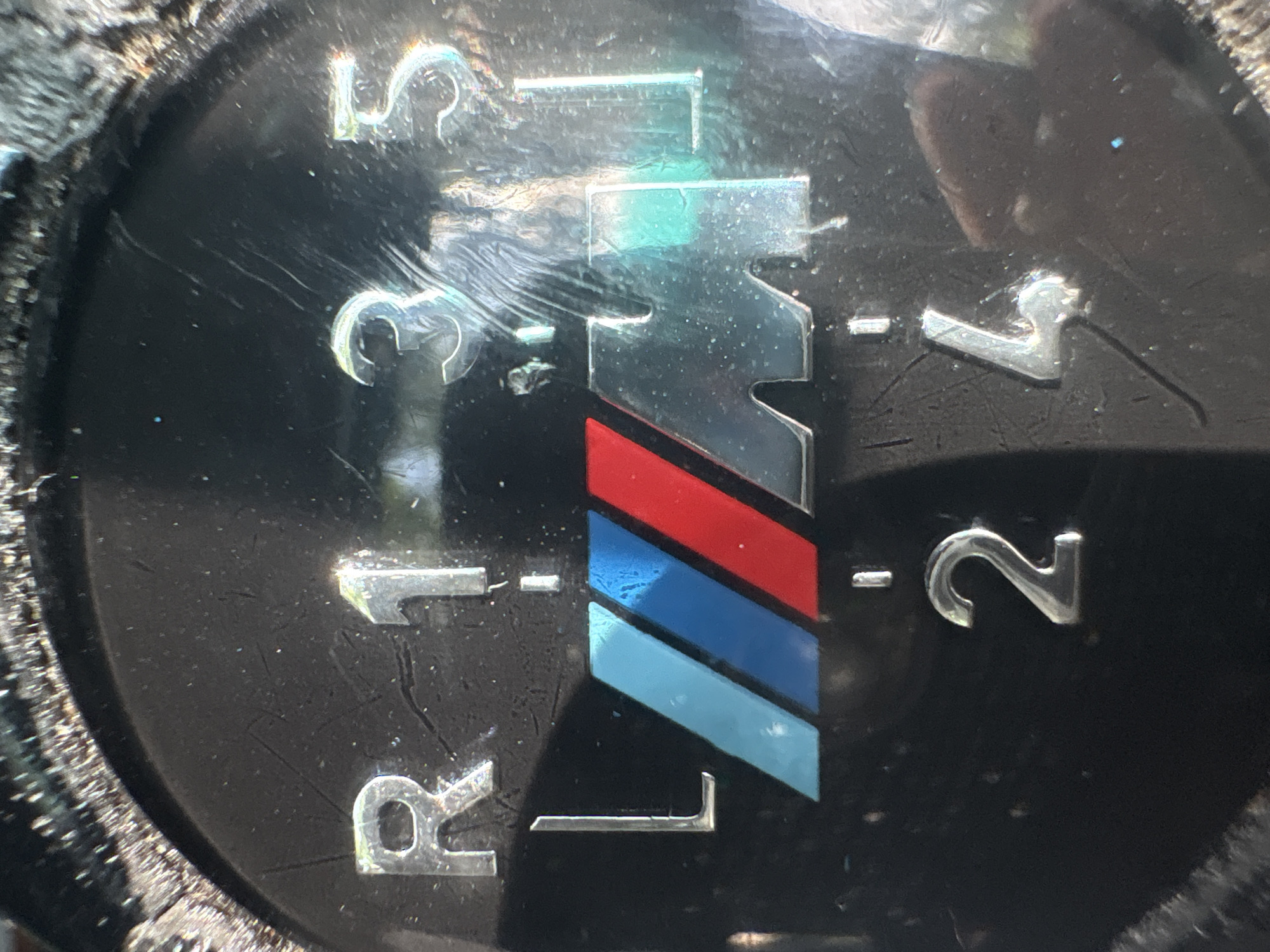 A close-up, high-angle shot captures a heavily worn, black gear shift knob from a manual transmission vehicle. The surface of the knob is severely scratched and scuffed, indicating significant use and age. Silver-colored numbers and letters display a 5-speed shift pattern: "R" (Reverse), "1", "2", "3", "4", and "5". The "R" is positioned at the top-left, while the forward gears follow a typical H-pattern, with 1, 3, and 5 in the upper row, and 2 and 4 in the lower row.
Dominating the center of the knob is a distinct "M" logo. This logo features a silver "M" symbol adjacent to three vertical stripes in descending order of color: red, dark blue, and light blue, characteristic of BMW's M performance division. Bright reflections, likely from a strong overhead light source, are visible on the glossy surface, creating highlights and minor visual distortion across the numbers, letters, and the "M" logo. The material surrounding the central insert of the knob appears textured and somewhat frayed, further emphasizing its used condition. The image provides a detailed view of a specific component from what is likely an enthusiast's vehicle.