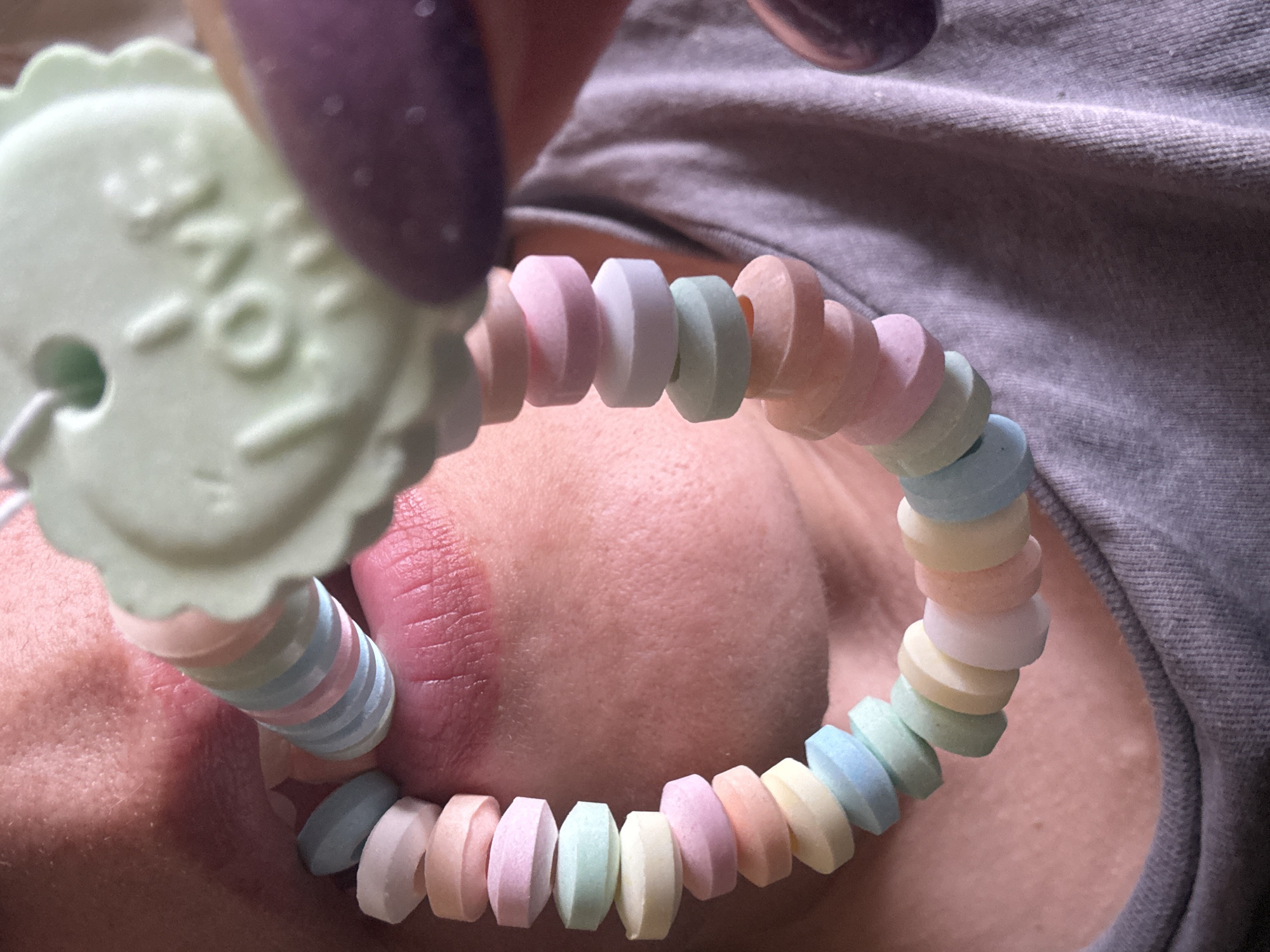 A close-up image shows a person holding a candy necklace. The necklace, made of pastel-colored, round candies, is stretched across the person's lower face, covering part of their mouth and chin. In the foreground, a large, light green candy with the word "LOVE" on it is visible. The person's fingers with purple nail polish hold the candy. The person is wearing a gray shirt. The location is not identifiable as Dmitrov, Russia from the visual information provided.