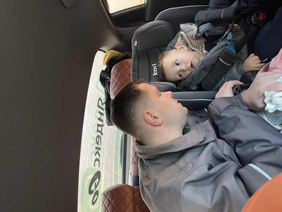 Inside a vehicle in Khimki, Russia, an adult male and a young child are observed in the rear passenger compartment. The adult male, with short hair and wearing a grey zip-up jacket, occupies the left rear passenger seat. He is turned towards the child, with a ring visible on his left ring finger, and his left hand is positioned near the child's lap, holding a white cloth. The child, seated to the right of the adult, is secured in a grey "kari" brand child safety seat. The child wears a striped shirt under a blue/grey puffer jacket and has short hair; their mouth is slightly open, and their eyes are wide, directed upwards and to the left. A colorful object hangs from the child seat harness on the child's right side, and a pink garment is on the child's lap. The vehicle's interior features brown, diamond-quilted headrests for the rear seats and a grey ceiling. Items including a dark jacket and a yellow cloth are present on the rear parcel shelf. The rear window displays a reversed "Yandex Go" logo in green and white, indicating the vehicle's affiliation with the ride-sharing service, against an indistinct outdoor background.