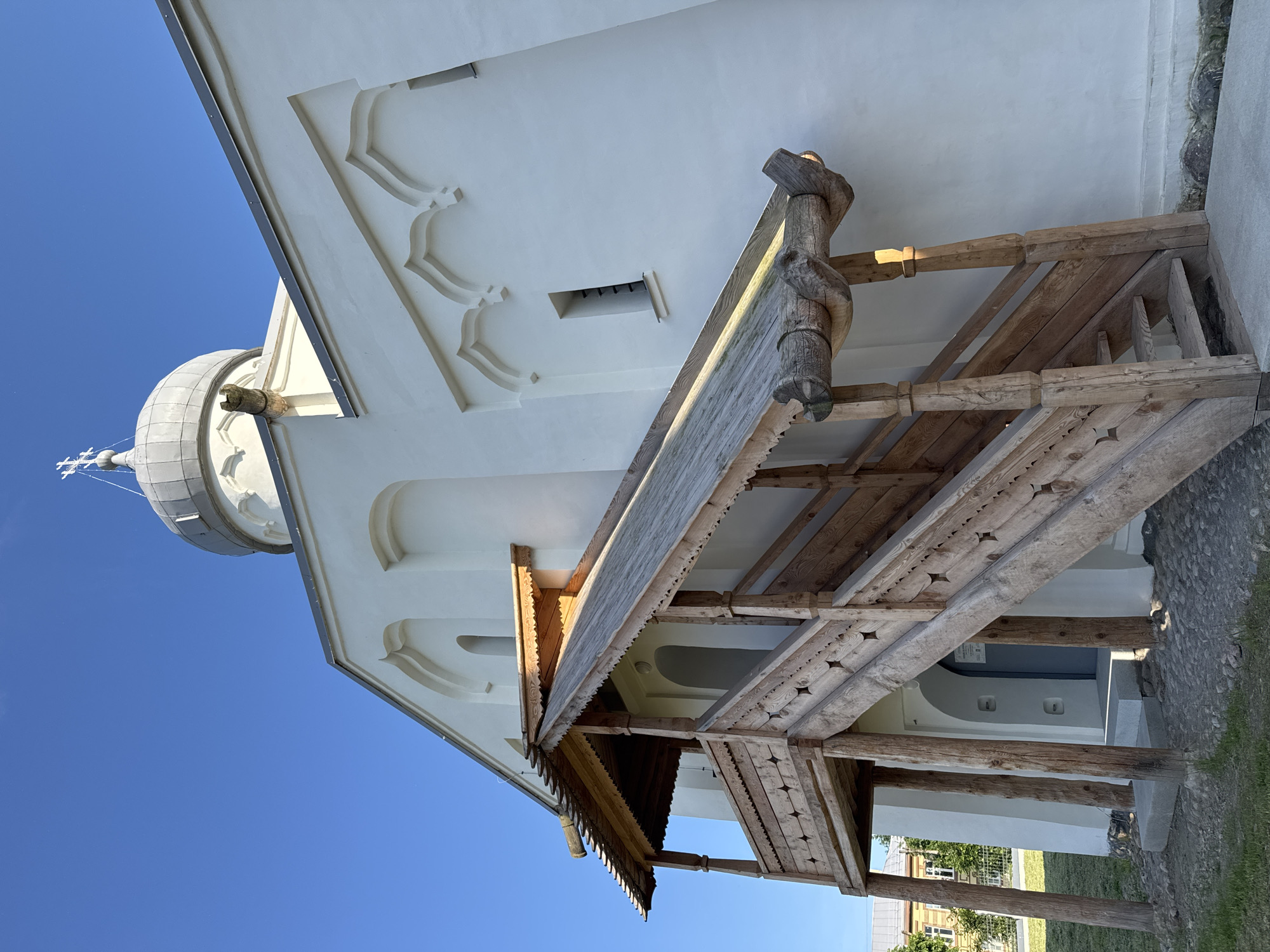 The photograph shows a side view of a white Orthodox church with a grey dome topped with a cross. The church has decorative arched elements above its windows. In the foreground, a wooden staircase with a covered walkway leads to an entrance. The staircase is elaborately carved with star patterns and is supported by thick wooden posts. The walkway is also made of wood with decorative trim. A stone gargoyle is visible near the dome. The ground around the church is covered with gravel and some grass. The sky is clear and blue. The architecture is characteristic of churches found in Veliky Novgorod, Russia. The image is captured from a low angle, looking up at the church.