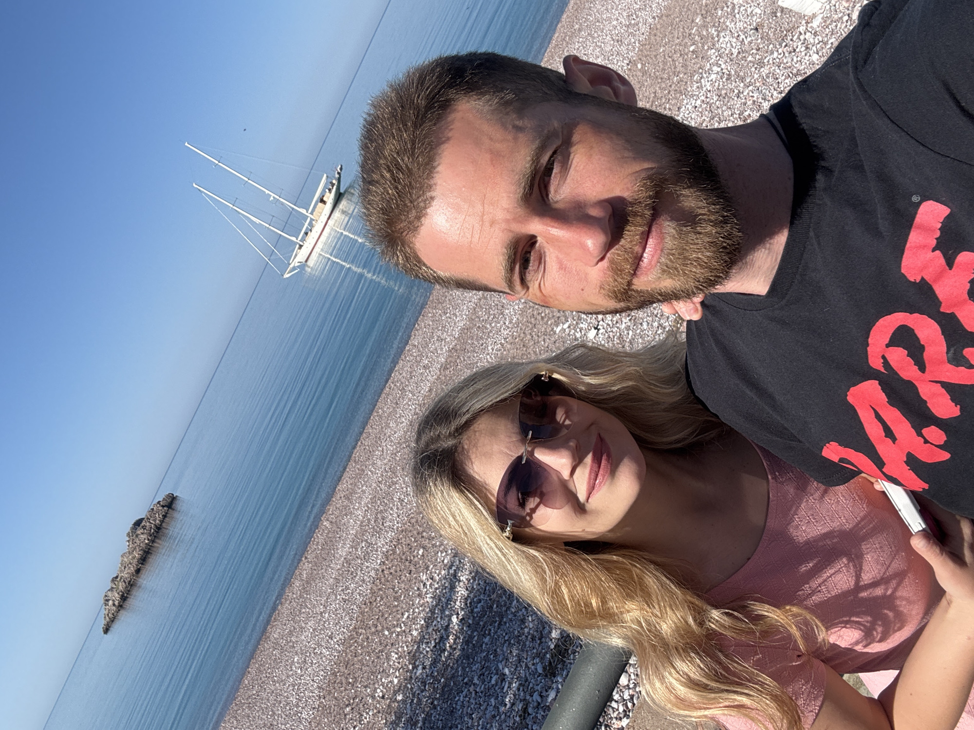 A brightly lit, landscape-oriented image captures a couple posing for a photo on a pebbly beach next to calm, clear blue water. The scene is set under a vast, clear blue sky.
In the foreground, slightly to the right, a man with short brown hair and a beard looks towards the viewer with a slight smile. He is wearing a black t-shirt with stylized red text on the front. To his lower left, partially behind him, a woman with long blonde hair and dark sunglasses is visible. She is wearing a light pink top and also looks towards the viewer with a subtle smile, holding a white object, possibly a phone, in her hand. The sunlight casts distinct shadows on their faces and clothing, indicating a sunny day.
The background reveals a serene marine environment. The tranquil blue sea extends to the horizon, meeting the clear sky. On the water in the middle distance, a white sailboat with two masts is visible. Further along the coastline, a small, elongated rocky islet protrudes from the water. The beach itself is covered in small, smooth pebbles.
The overall impression is one of relaxation and enjoyment during a sunny day by the sea, likely in Sveti Stefan, Montenegro, as suggested by the location context. The couple appears content, capturing a moment together amidst a picturesque coastal backdrop.